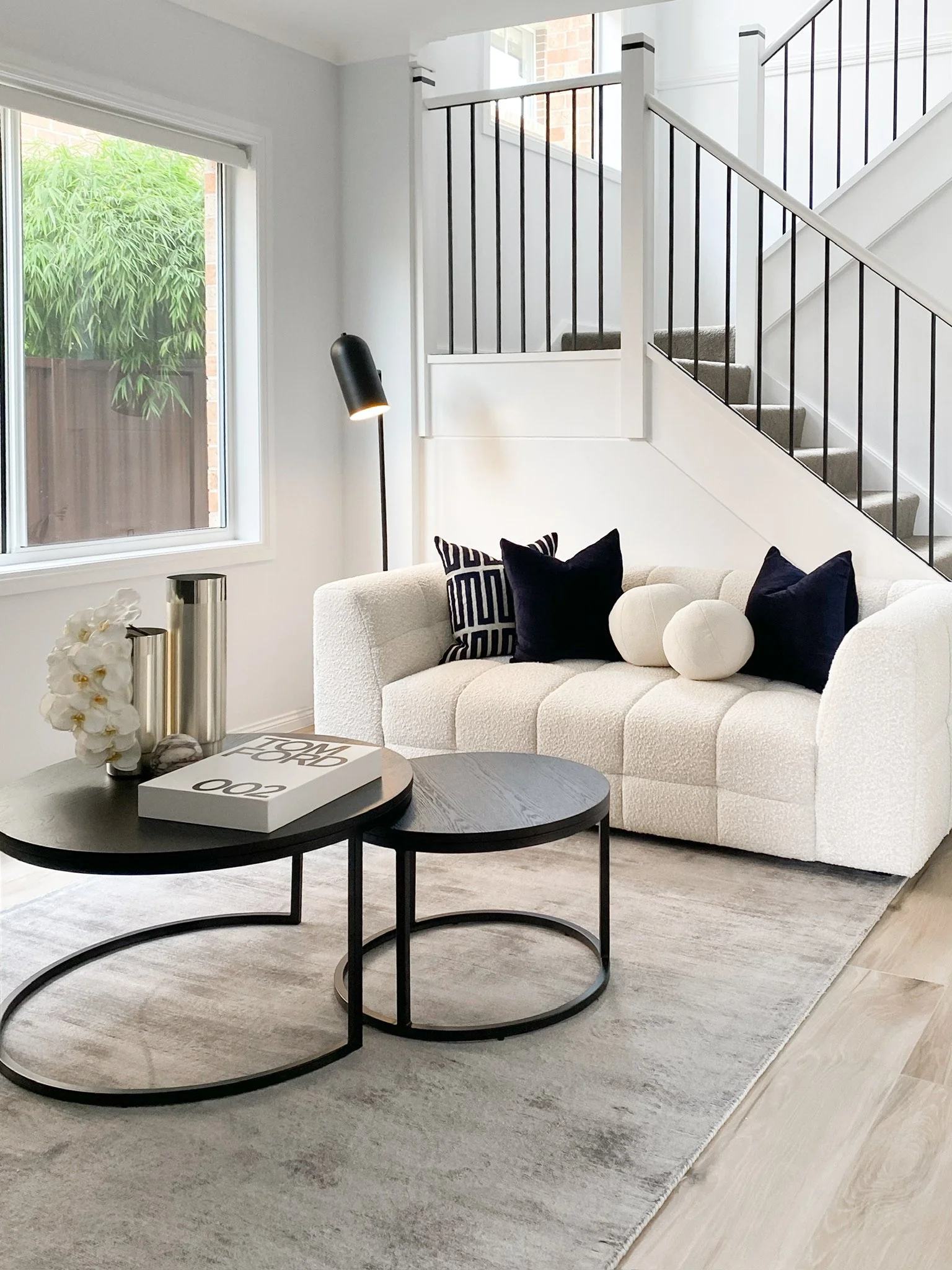 Modern living room with a white upholstered sofa, black and white pillows, and circular nesting coffee tables. A decorative book and a vase with white flowers sit on the tables. A minimalist floor lamp is next to the sofa, and a staircase with black railing is in the background.