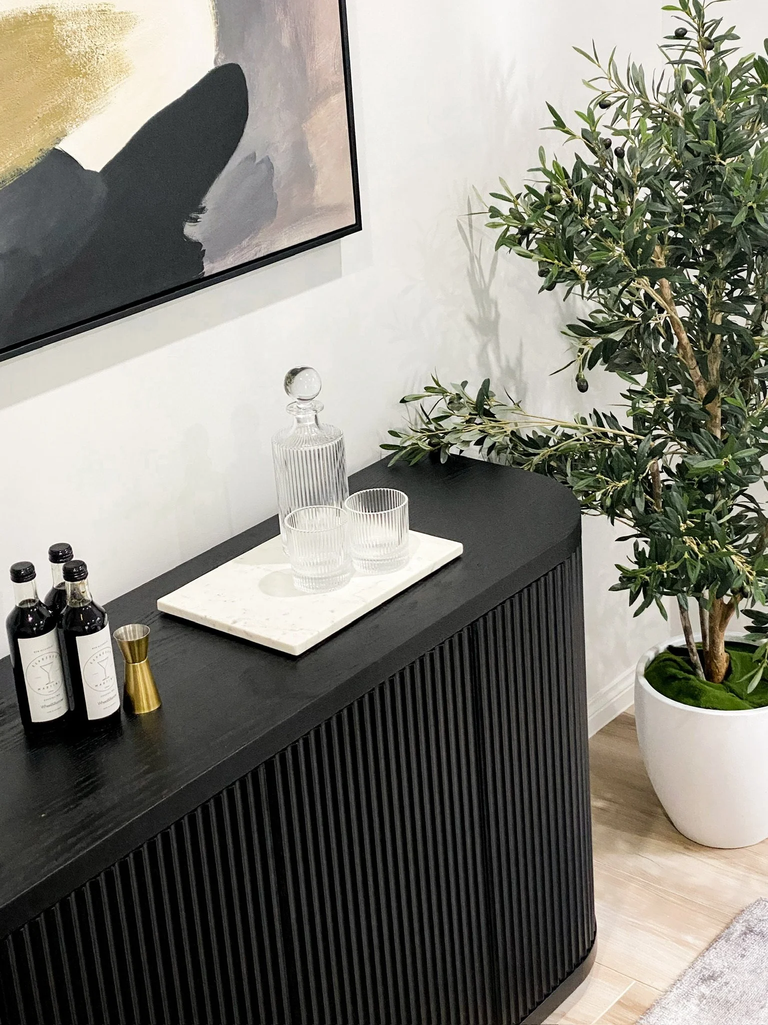 Modern black sideboard with abstract painting, glass decanter, two cups, bottles, jigger, and potted plant.