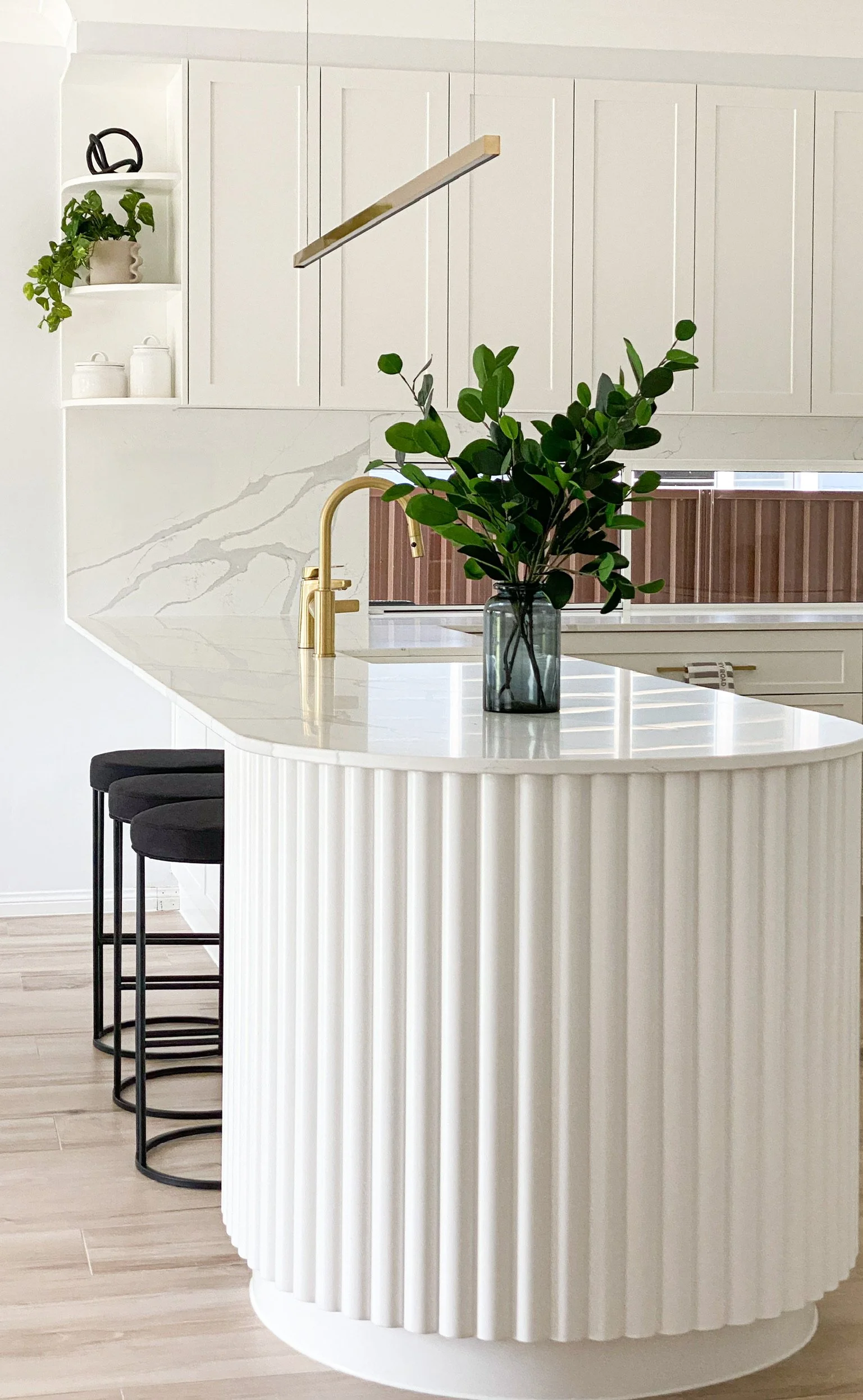 Modern kitchen with a white fluted island countertop, gold faucet, green plant in a vase, black stools, white cabinets, and decorative shelves.
