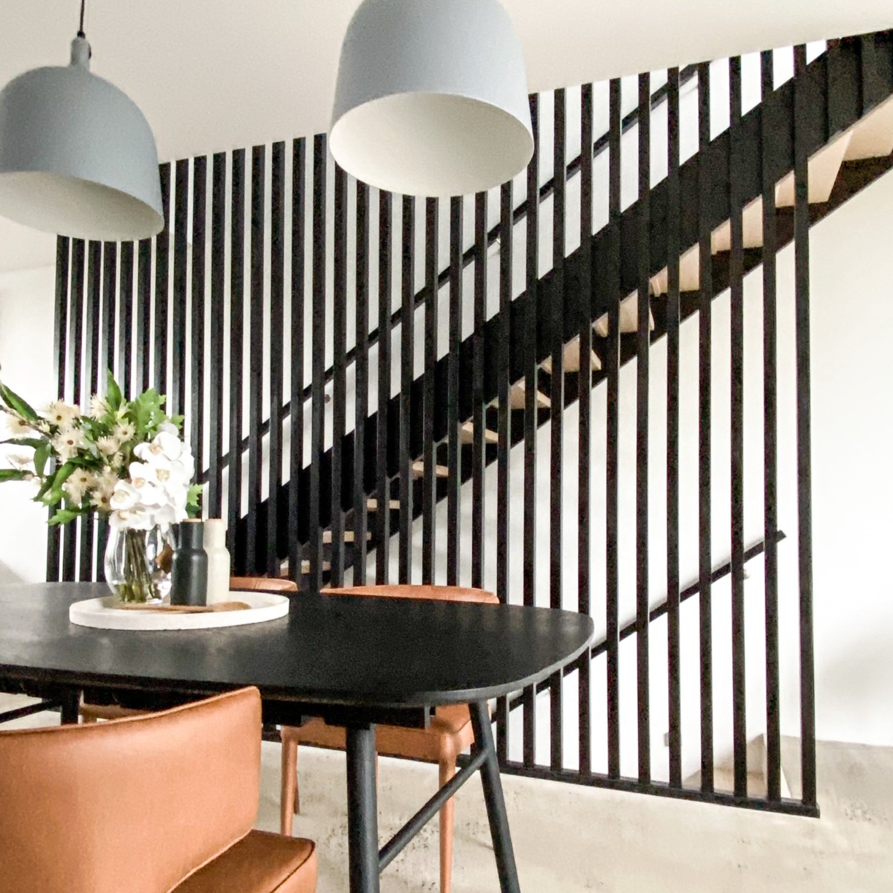 Modern dining area with black table, brown chairs, and stylish staircase with vertical wooden slats. Decor includes two pendant lights and a vase with flowers.