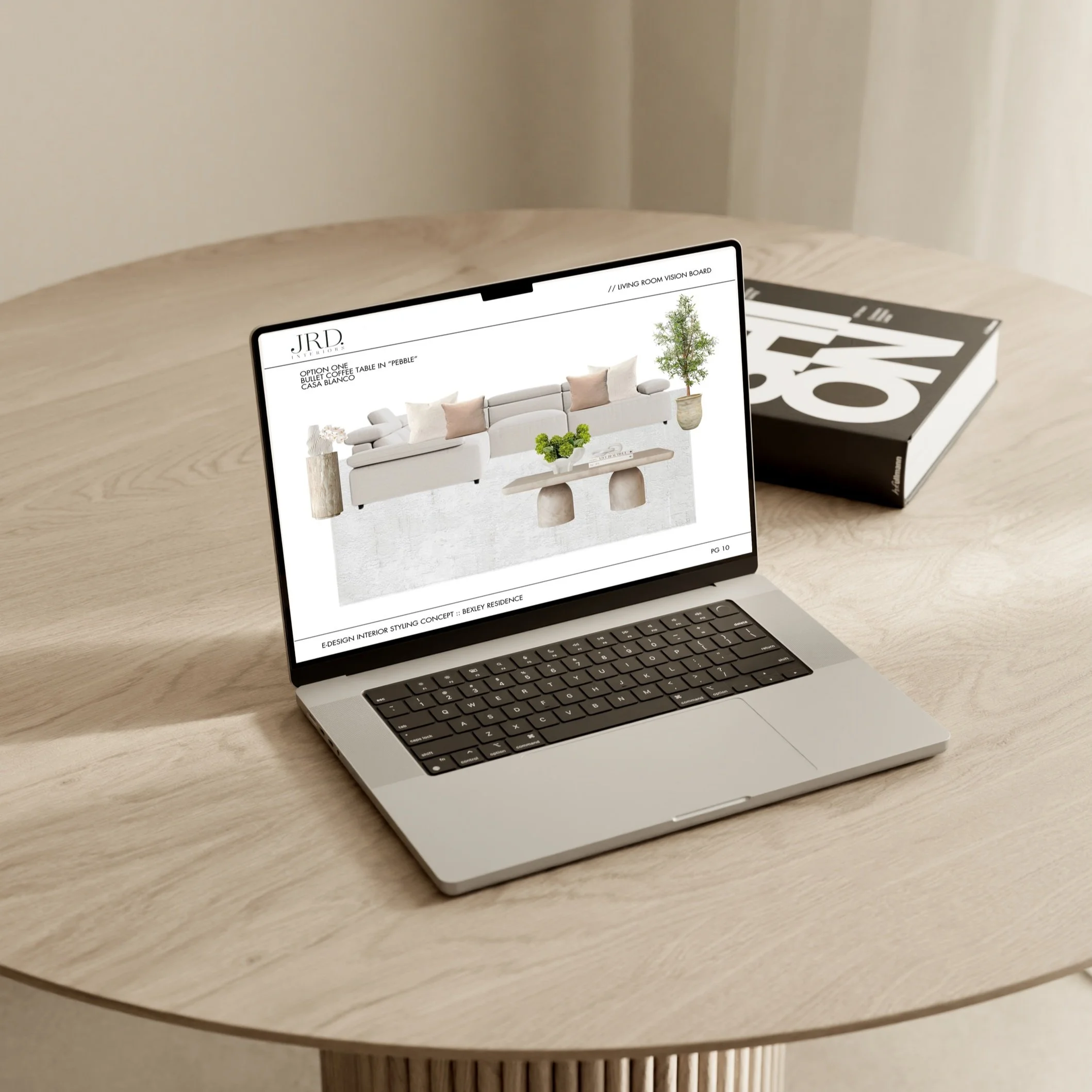 Laptop on a wood table displaying interior design mockup with sofa, pillows, and plants, next to a closed book.