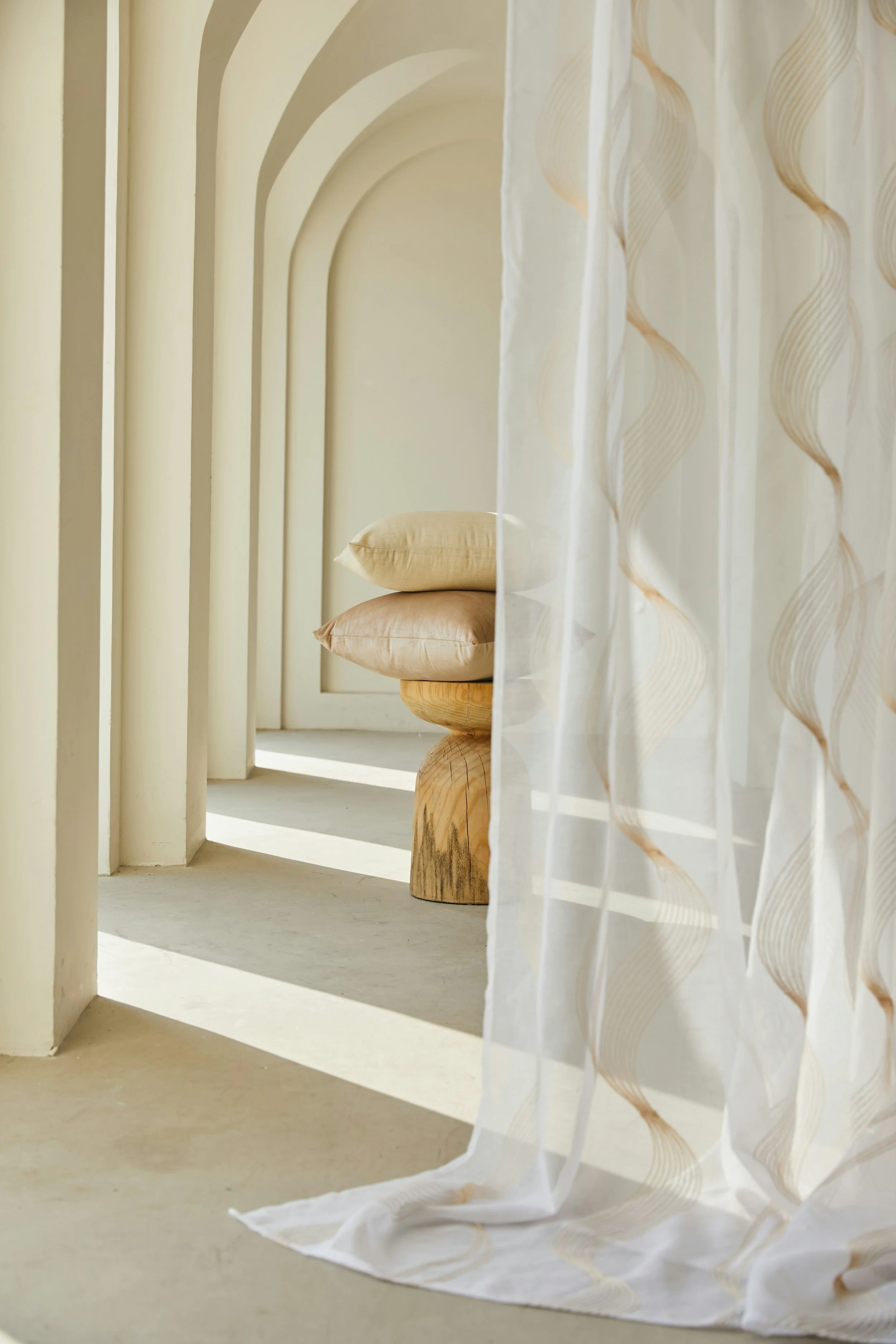 White curtain with wave patterns partially open, revealing two beige cushions stacked on a wooden stool in a hallway with arched openings and soft lighting.