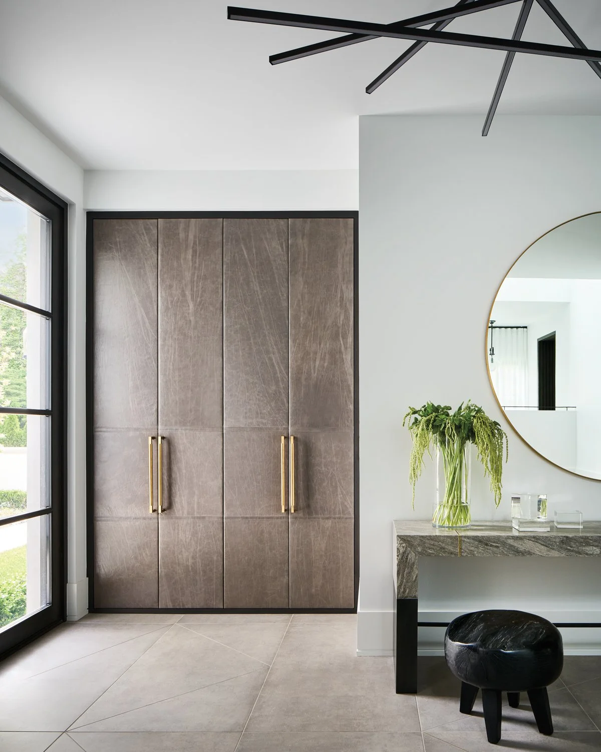 Modern entryway with large wooden doors, black chandelier, round mirror, stone console table, black stool, and a glass vase with greenery.