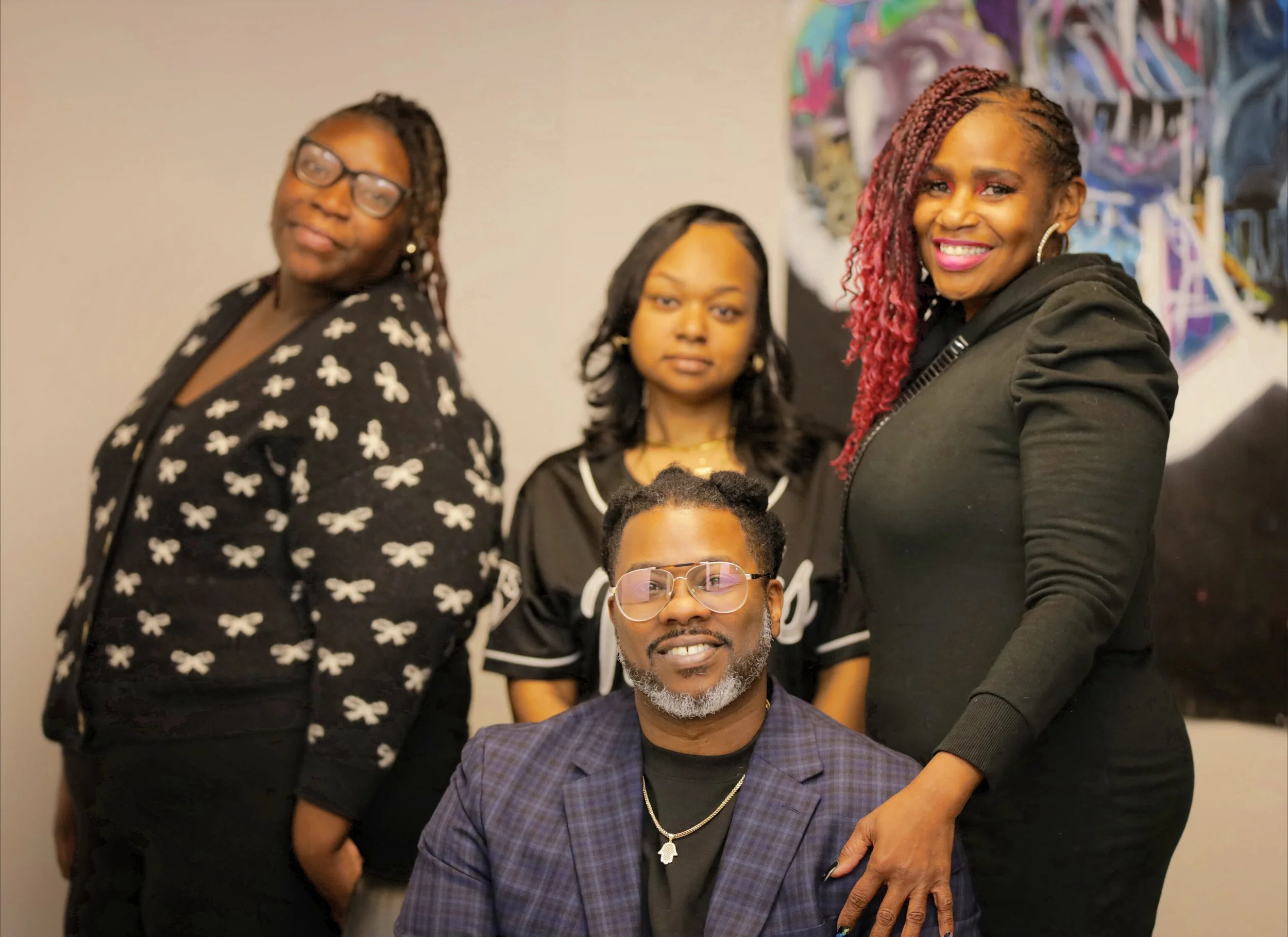 Four diverse adults posing indoors; a man seated in front with glasses and a plaid blazer, three women behind him with varying hairstyles and expressions, all dressed casually with artistic background.