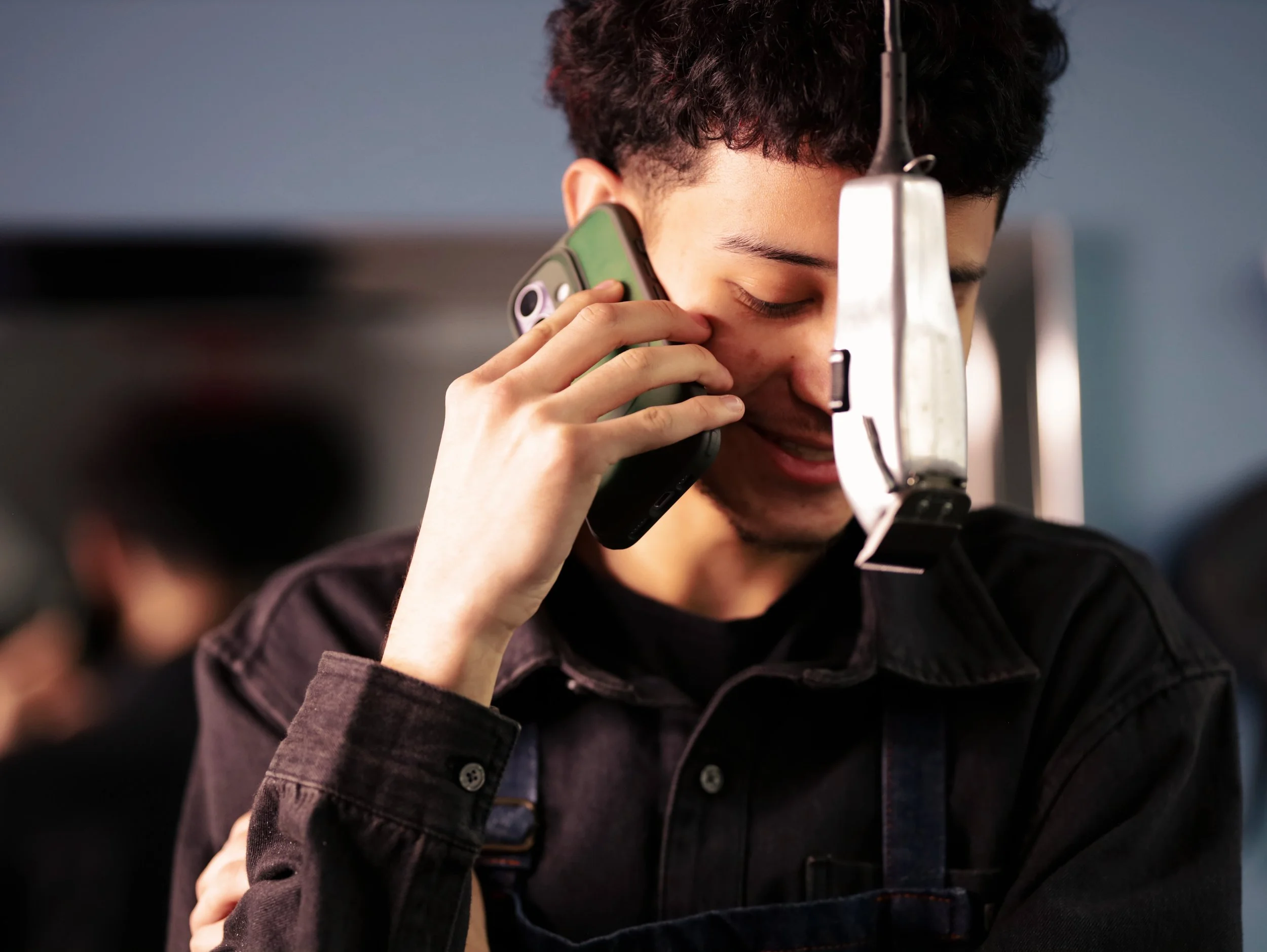 A young man working at Aqua cuts Barbershop with curly hair, smiling with eyes closed, talking on a phone. He is wearing a black jacket and has a hair clip in his hair, with a blurred background.
