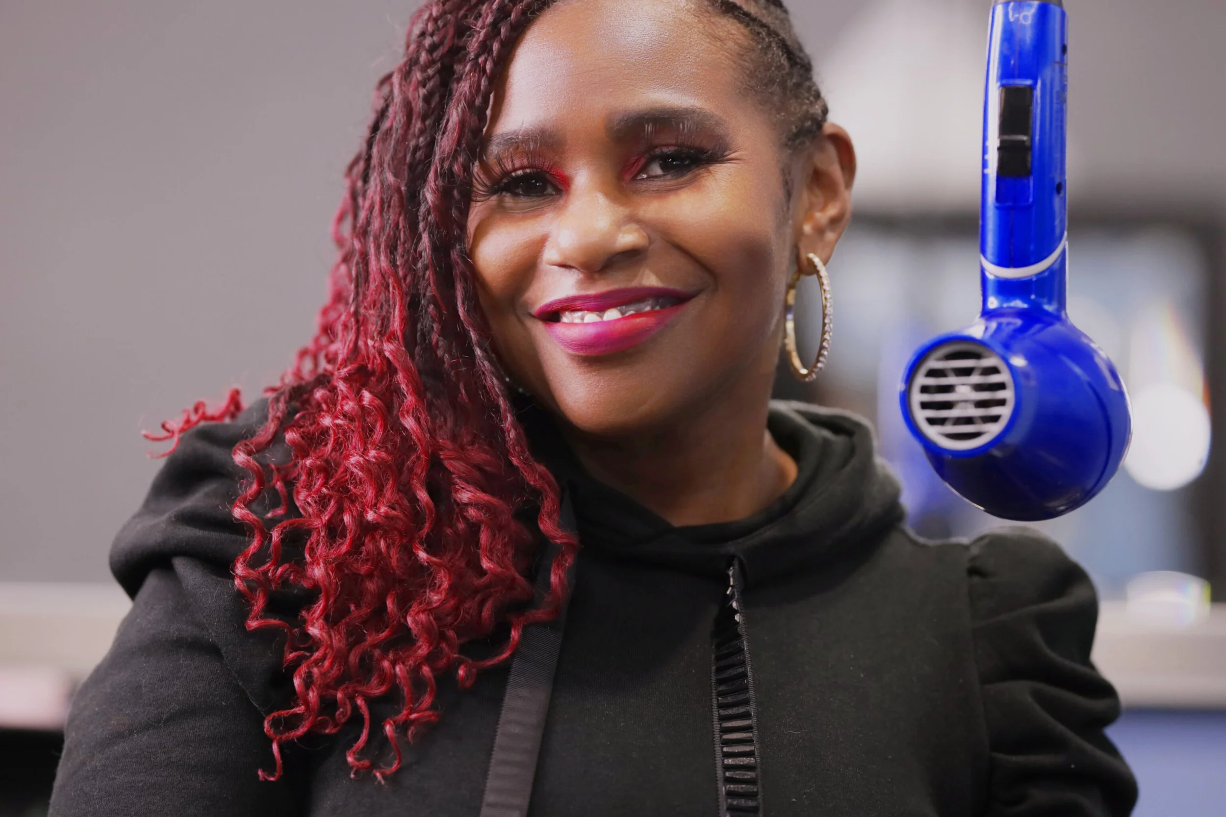 A woman with red and purple curly hair working at Aqua Cuts Barbershop smiling in front of a blue hair-dryer, wearing a black top and hoop earrings.