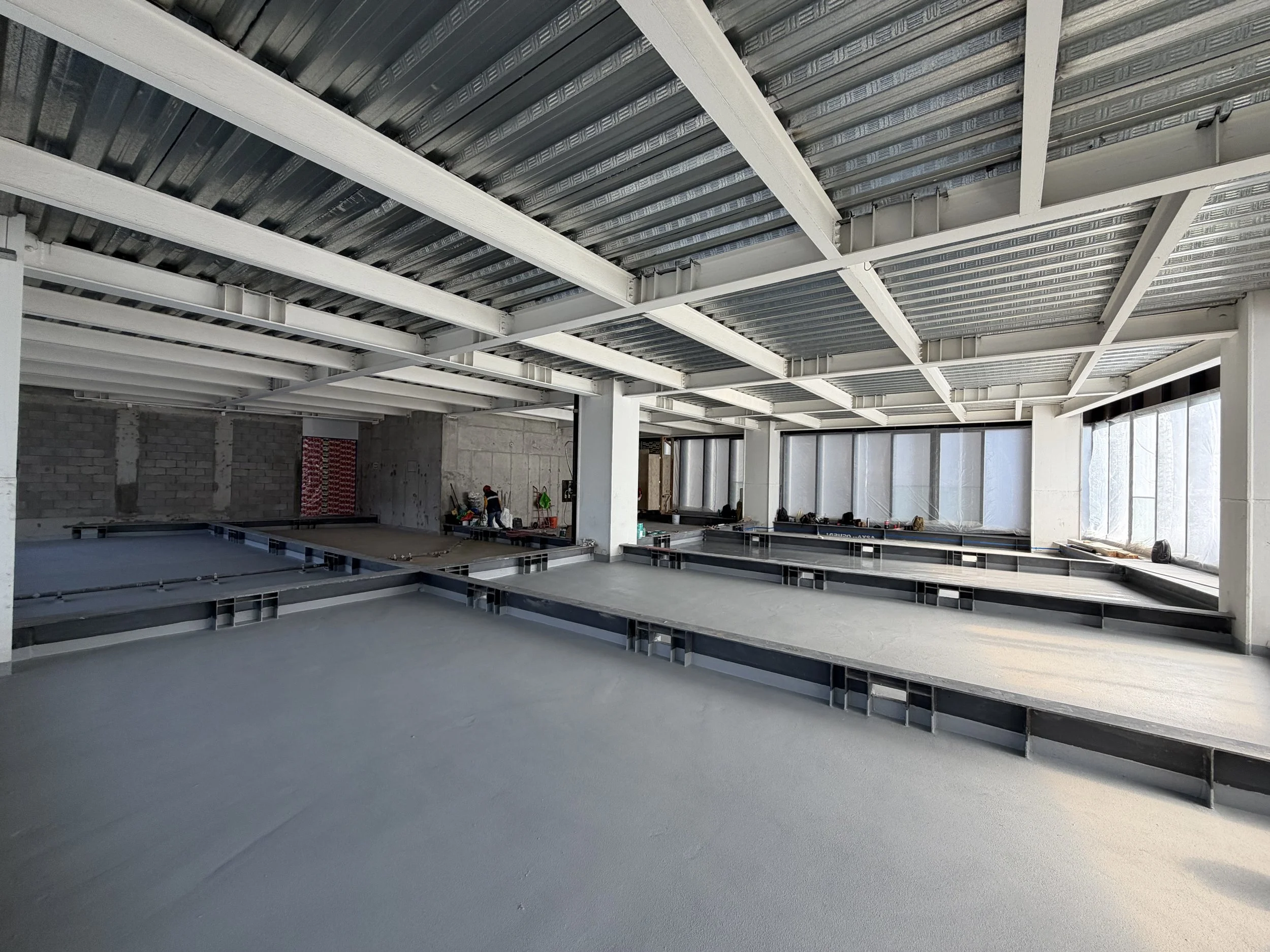 Piso de concreto en construcción, dividido en dos secciones con diferentes acabados, en un espacio interior con paredes de bloques de cemento y ventanas con plástico transparente.