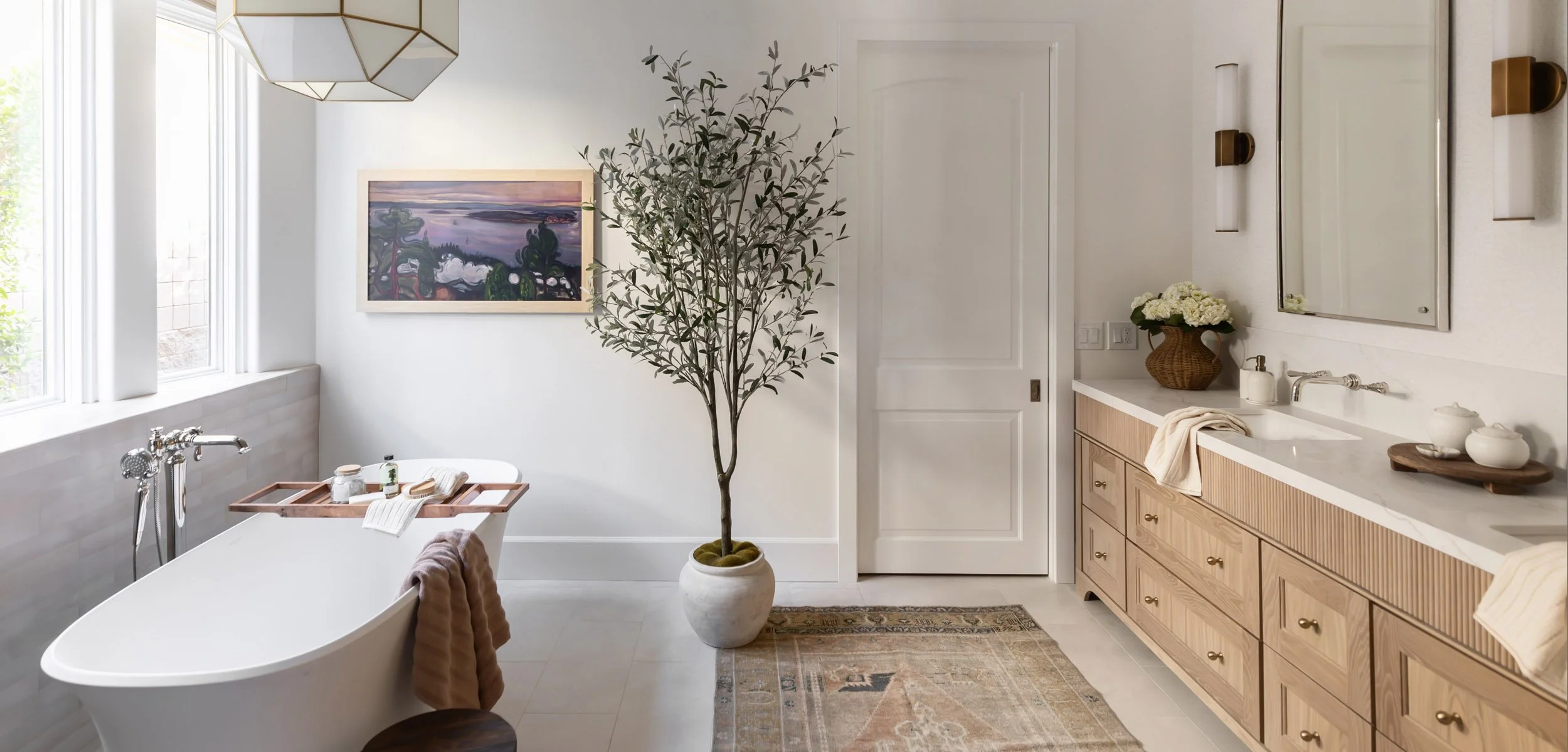 Primary bathroom in a Scottsdale home with freestanding soaking tub, zellige-style tile, potted olive tree, vintage-style rug, and light oak double vanity with brass and nickel fixtures
