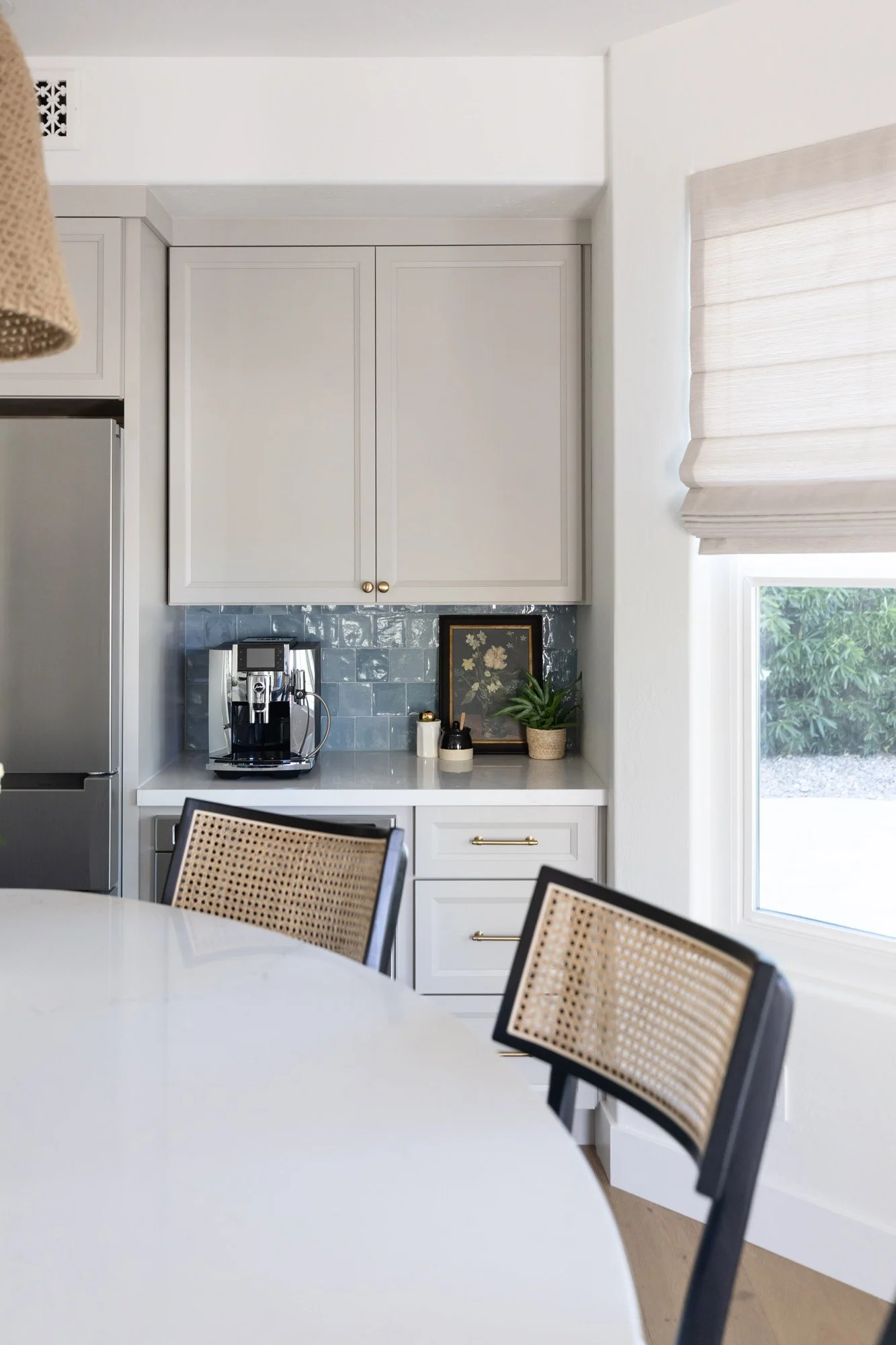 Kitchen corner with white shaker cabinetry, blue zellige tile backsplash, gold hardware, cane bar stool, stainless steel fridge, and woven roman blind