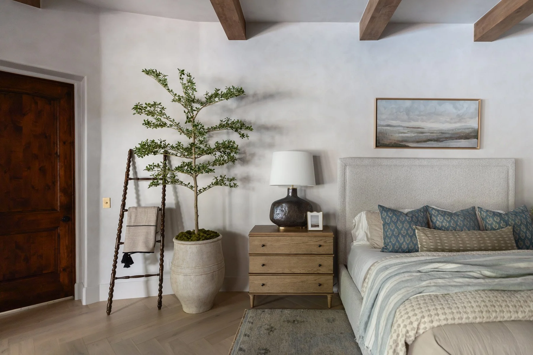 Primary bedroom with iron bed frame, upholstered bedhead, blue accent pillows, oak dresser, black ceramic lamp, ladder shelf, and large potted tree beside dark wood door
