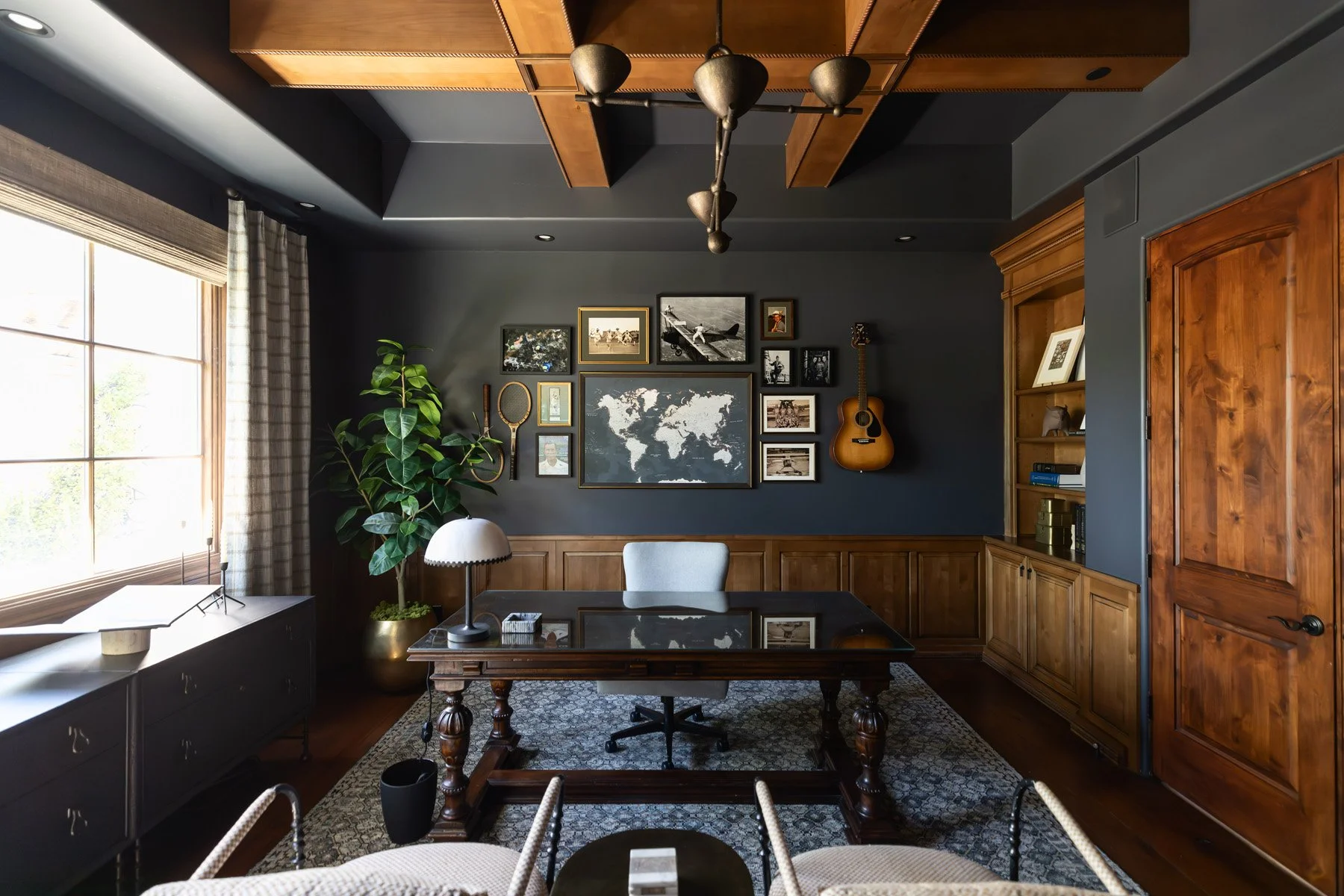 Dark home office with navy walls, dark wood wainscoting, ornate writing desk, framed photo gallery, guitar, world map artwork, and window with linen drapery