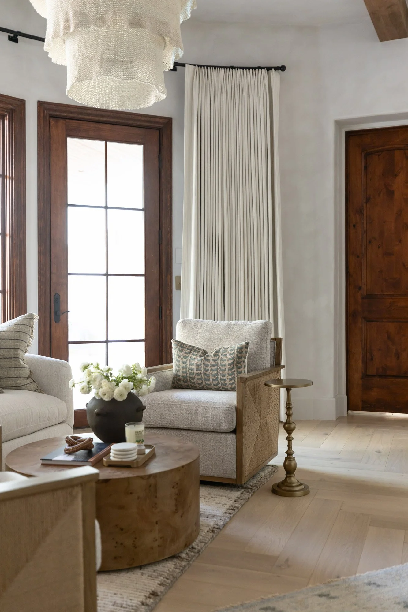 Sitting room with white armchair, round stone coffee table, brass candlestick, white floral arrangement, and dark wood French door with neutral drapery