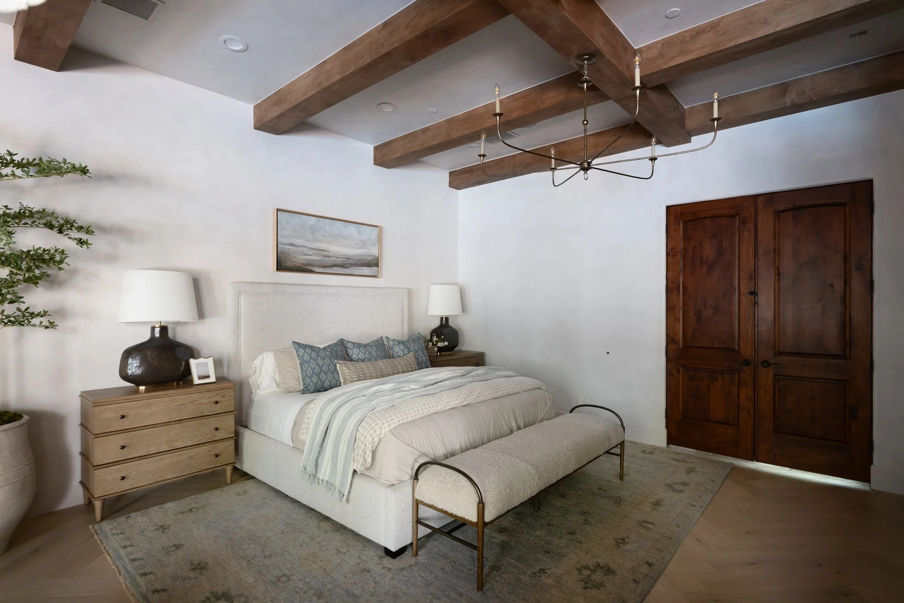 Primary bedroom with iron bed frame, upholstered bedhead, oak dresser, black ceramic lamp, dark wood double doors, and exposed wood ceiling beams