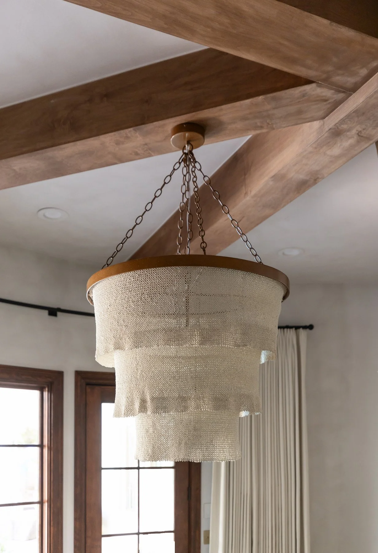 Woven fringe drum pendant light hanging from exposed wood ceiling beams with dark wood French windows behind