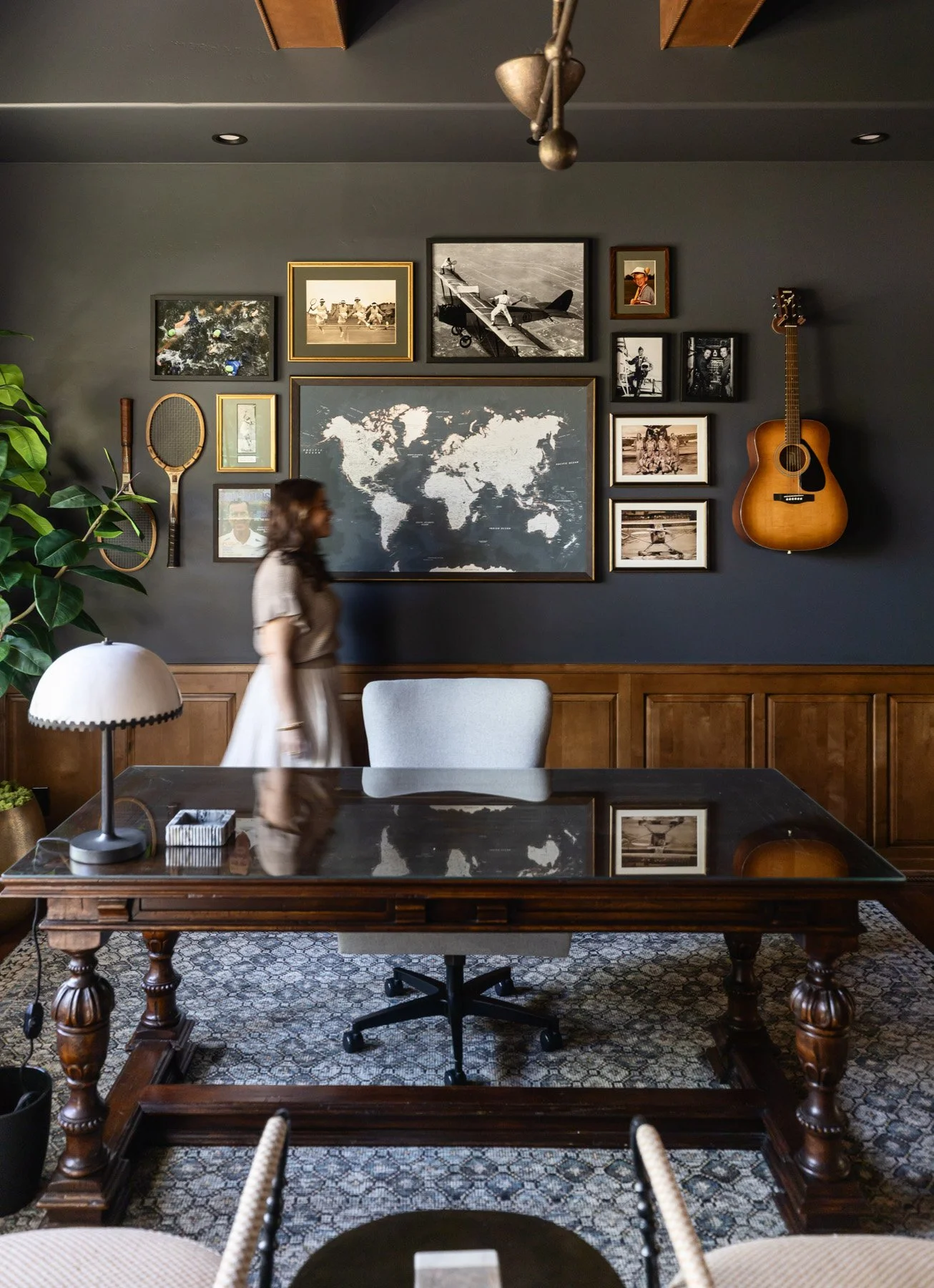 Dark home office with navy walls, ornate writing desk, grey upholstered chair, world map artwork, vintage framed photo gallery, guitar, tennis rackets, and dark wood wainscoting
