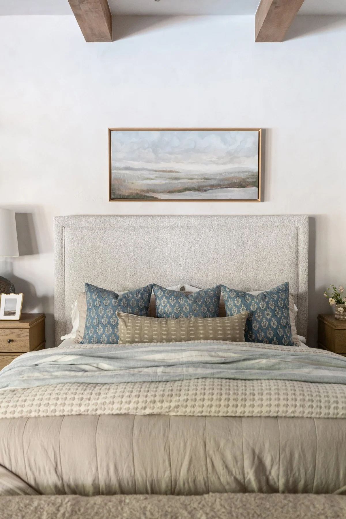 Primary bed with upholstered headboard, blue patterned accent pillows, and layered neutral checked and striped bedding with landscape artwork above