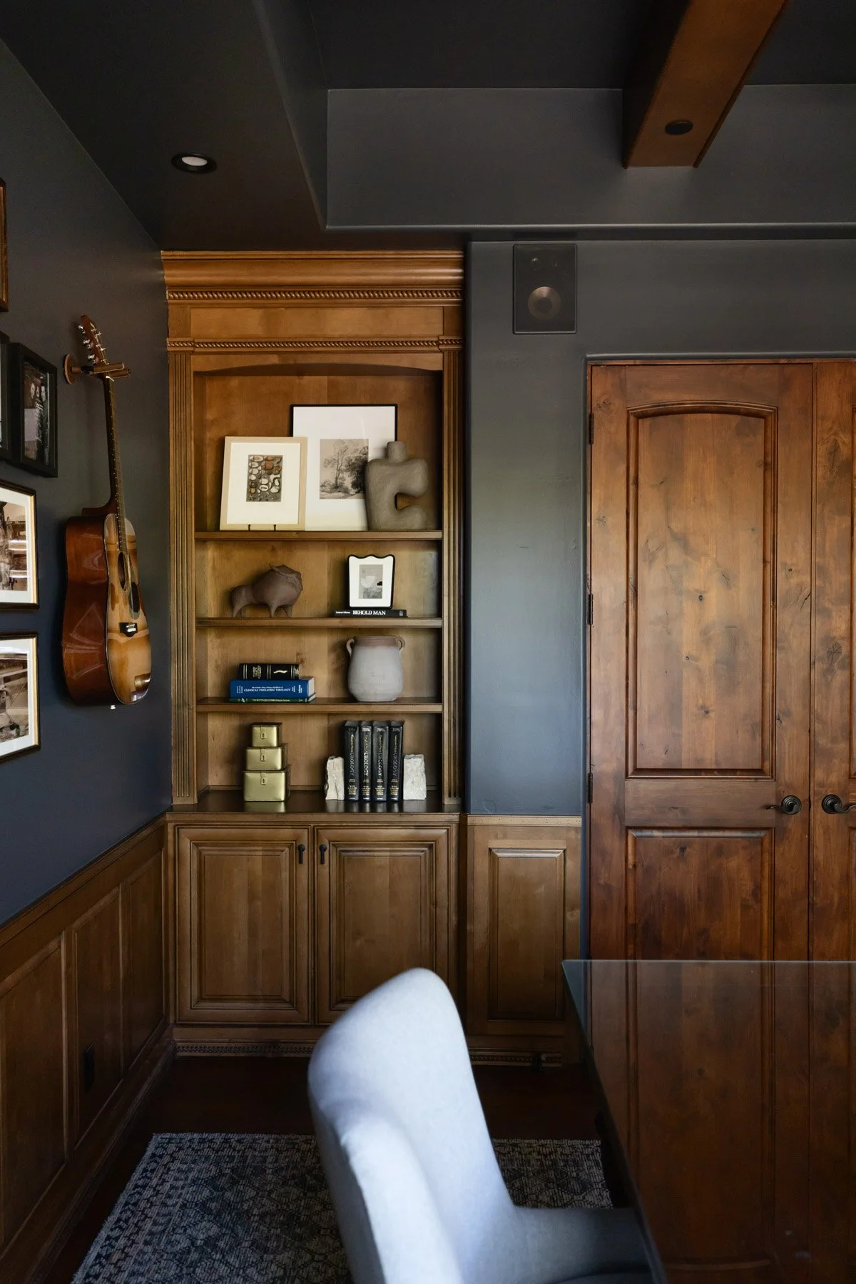 Dark wood built-in cabinetry with arched detail, open shelving with guitar and decorative objects, and white upholstered chair beside dark wood door