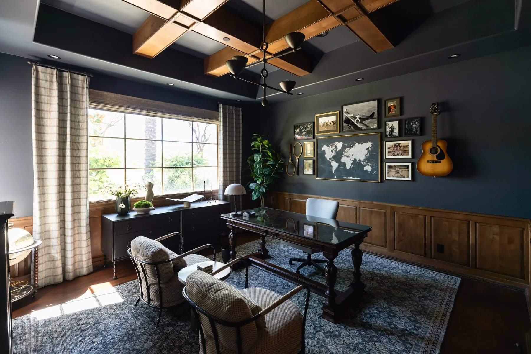 Dark home office with navy walls, dark wood wainscoting, ornate writing desk, upholstered chair, world map artwork, guitar, framed photo gallery, and large window with linen drapery