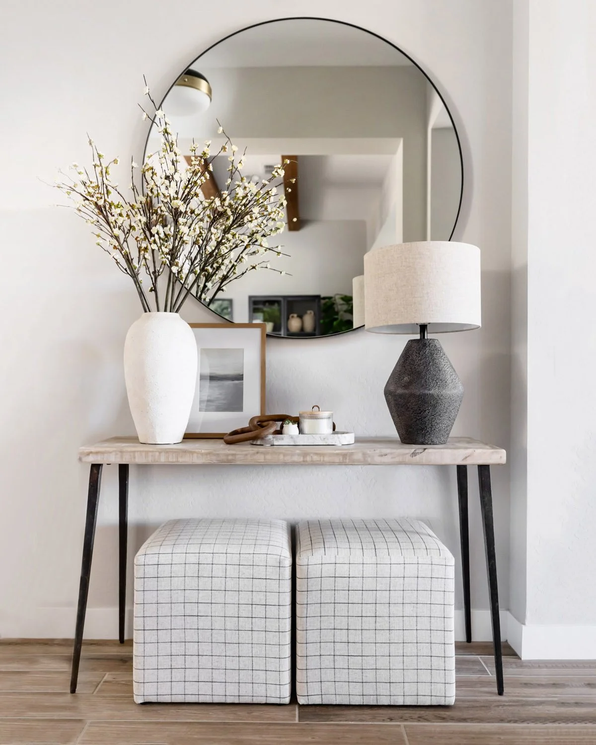 Entryway console in a Phoenix home with black iron legs, white ceramic vase with dried branches, round mirror, table lamp, and grid upholstered ottomans below