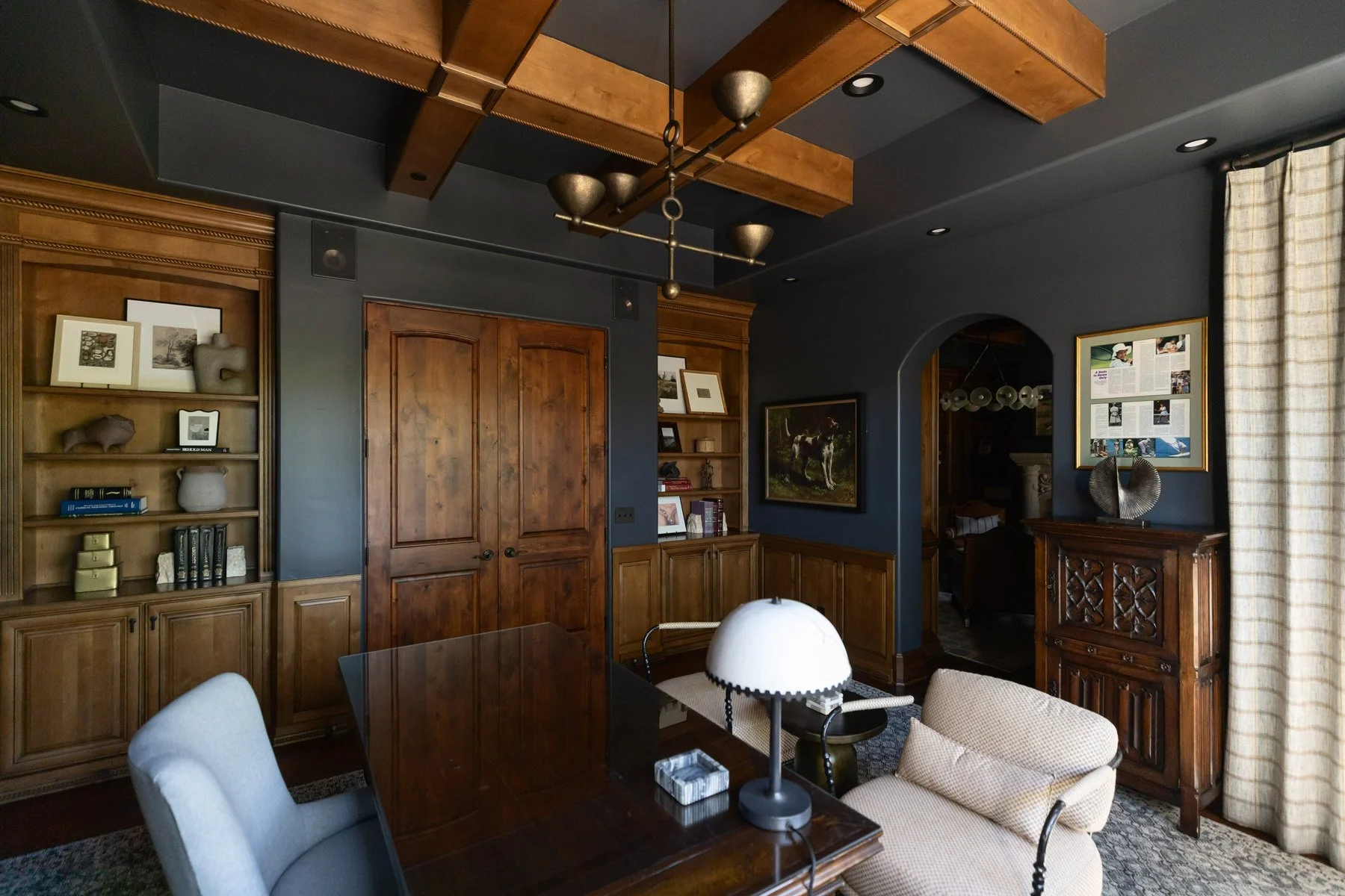 Dark home office with navy walls, dark wood wainscoting, writing desk, white table lamp, built-in cabinetry with arched detail, exposed wood ceiling beams, and upholstered sofa
