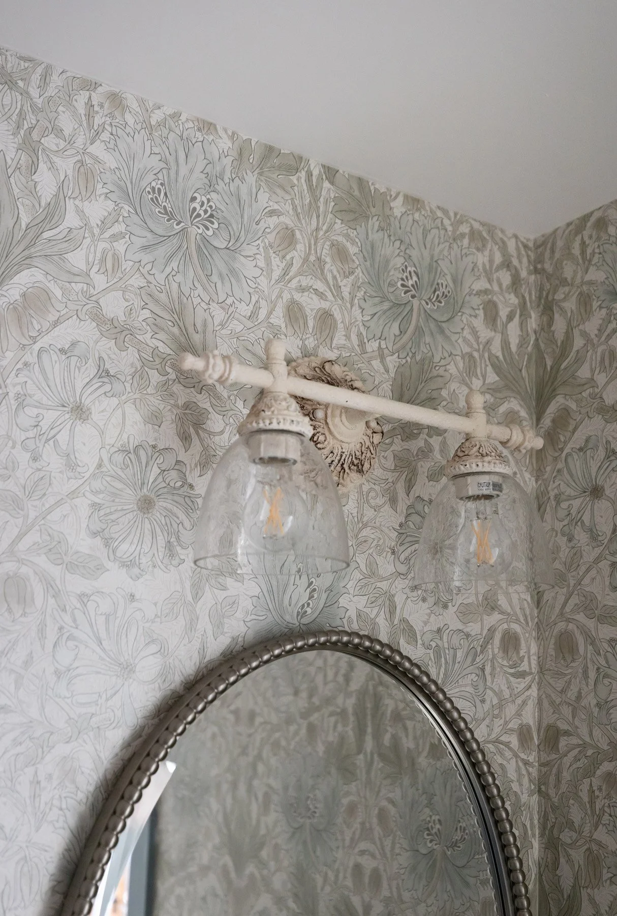Bright bathroom vanity light detail with white double sconce bar, botanical wallpaper, and oval mirror edge below