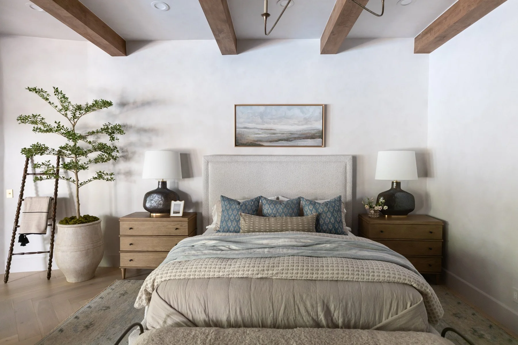 Primary bedroom with upholstered bedhead, blue accent pillows, layered neutral bedding, oak dresser, ladder shelf, potted tree, and landscape artwork