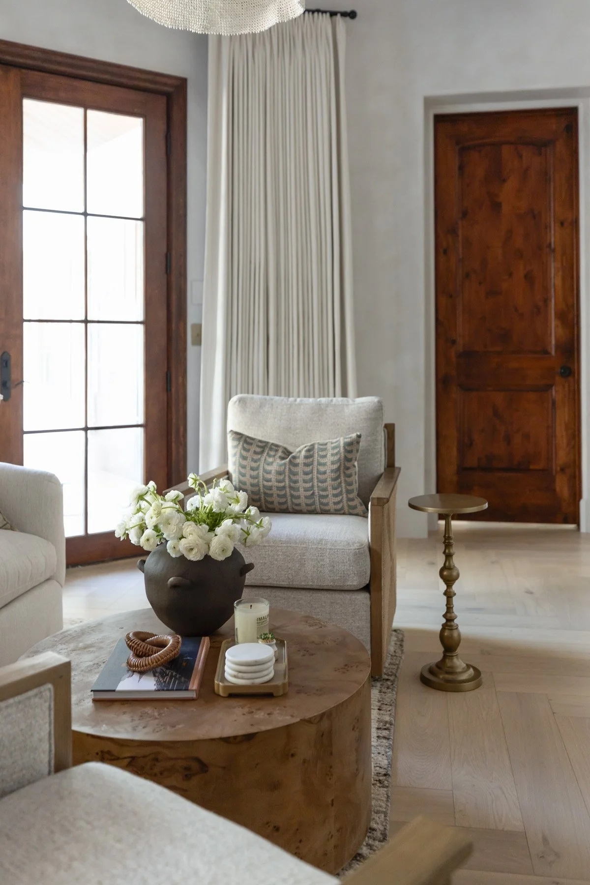 Sitting room with white armchair, round coffee table, brass candlestick, white floral arrangement, neutral drapery, and dark wood French doors