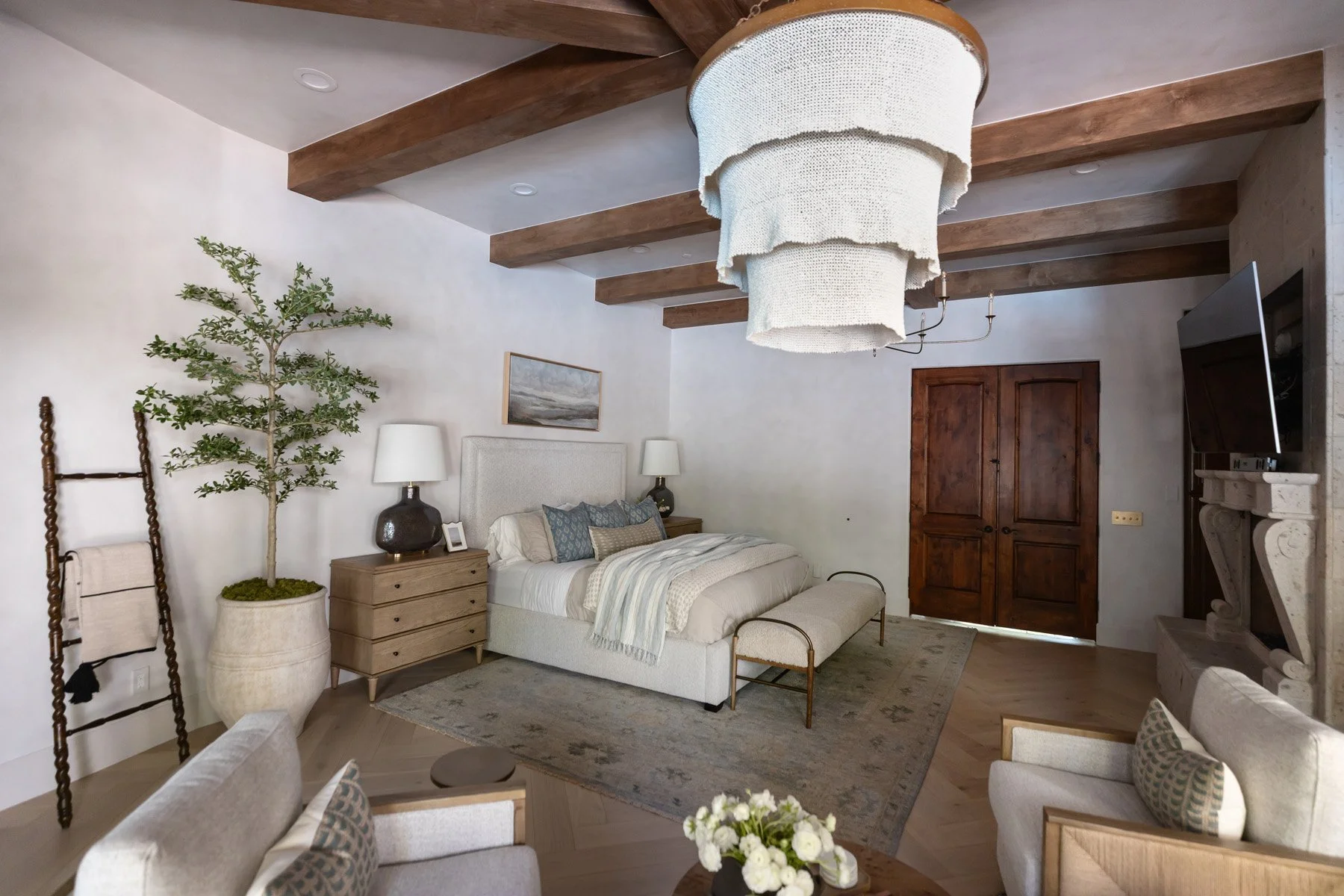Primary suite with iron bed frame, oak dresser, ladder shelf, potted tree, drum pendant light, dark wood doors, and exposed wood ceiling beams