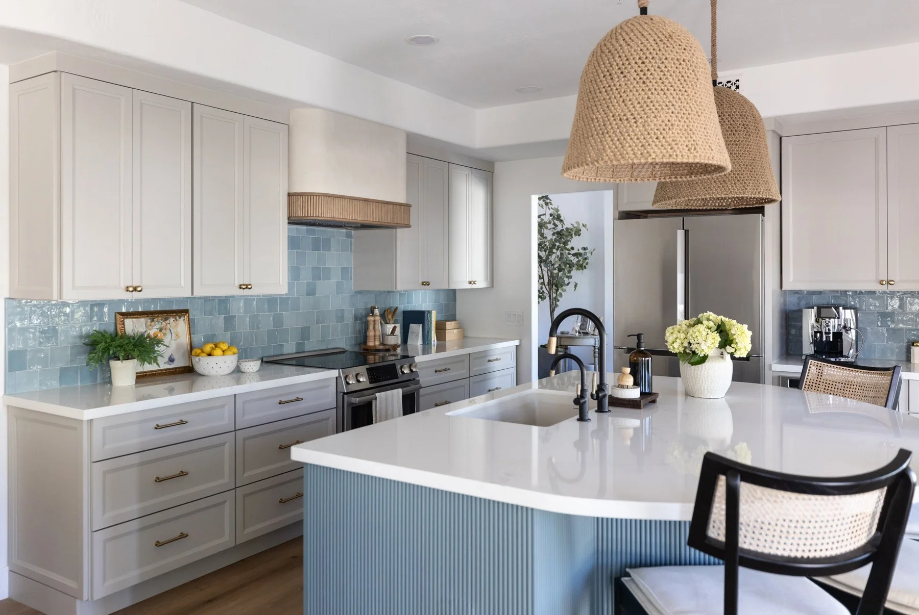 Kitchen overview with white shaker cabinetry, blue zellige tile backsplash, woven pendant light, blue fluted island with white quartz countertop, black cane bar stool, and yellow floral arrangement