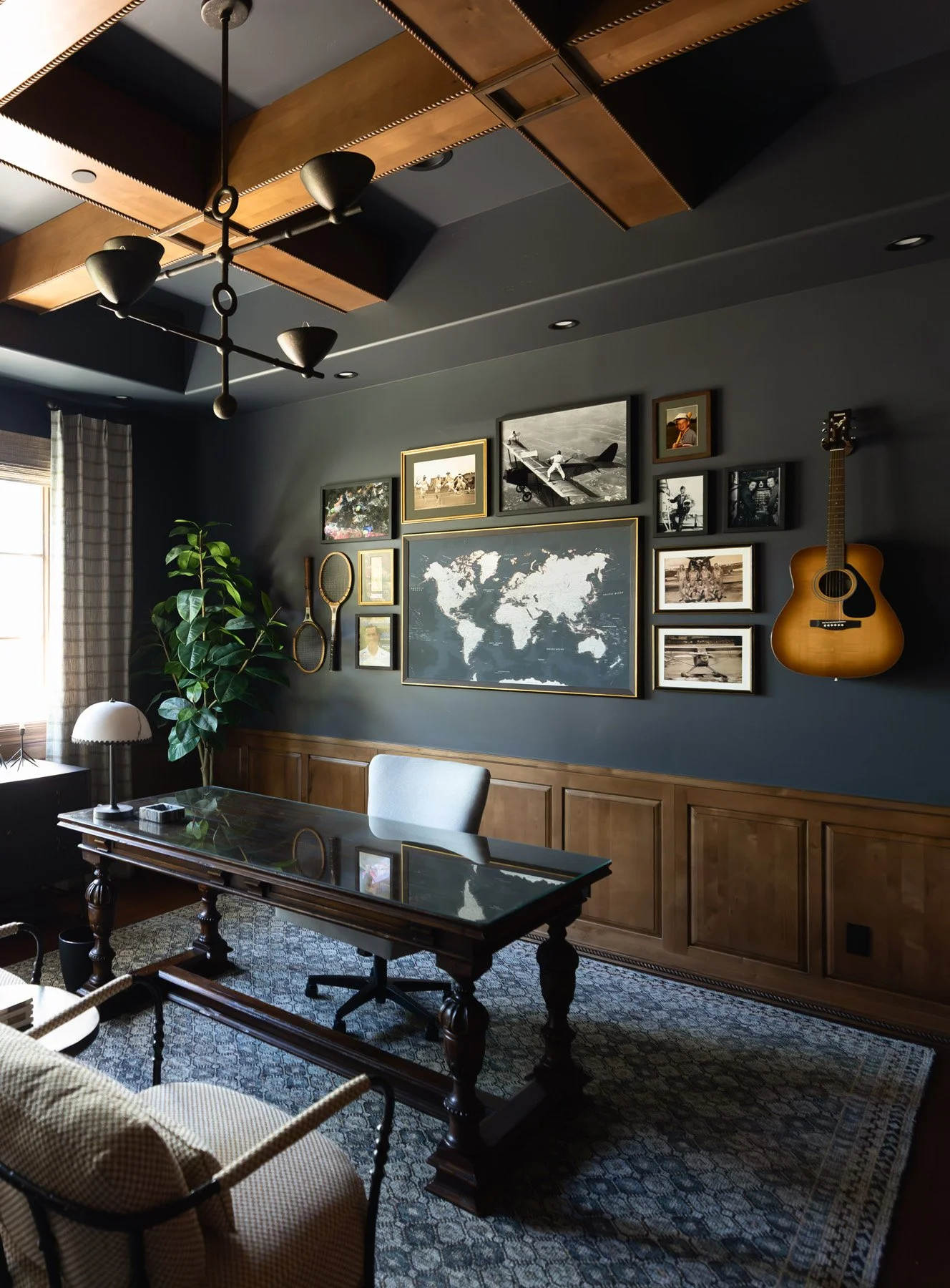 Dark home office with navy walls, dark wood wainscoting, ornate writing desk, world map artwork, guitar, framed photo gallery, and exposed wood ceiling beams
