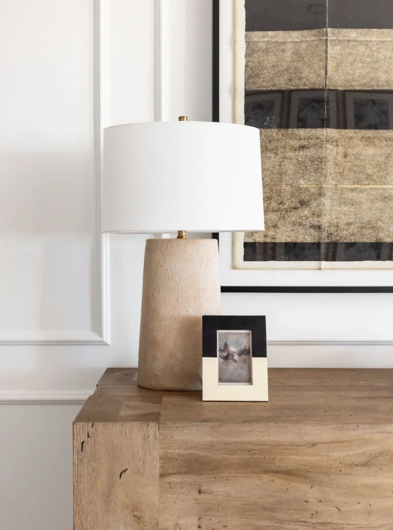 Stone textured table lamp with white linen shade and brass detail on a reclaimed wood console, with a small black and white framed photo and large abstract artwork behind