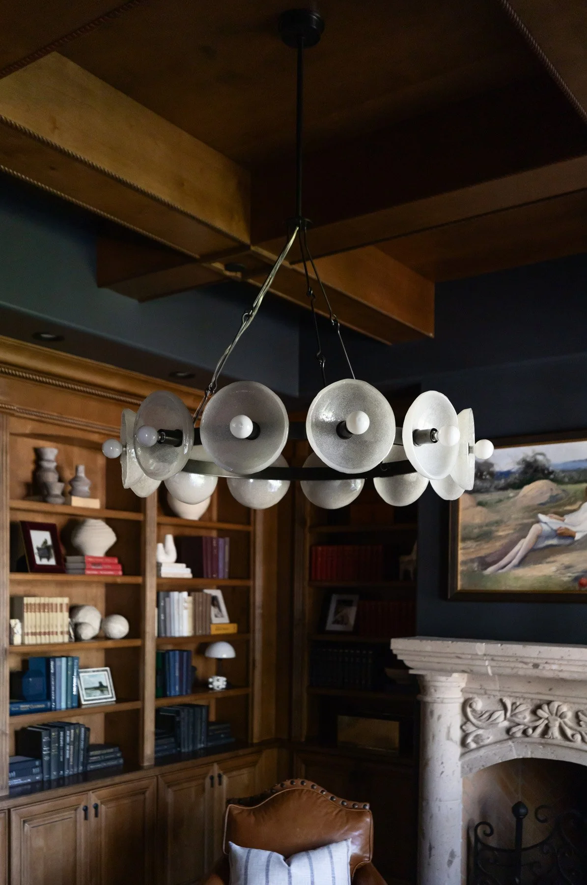 Dark study with ornate stone fireplace, leather armchairs, sculptural pendant light, dark wood built-in bookshelves, and framed landscape artwork above mantle