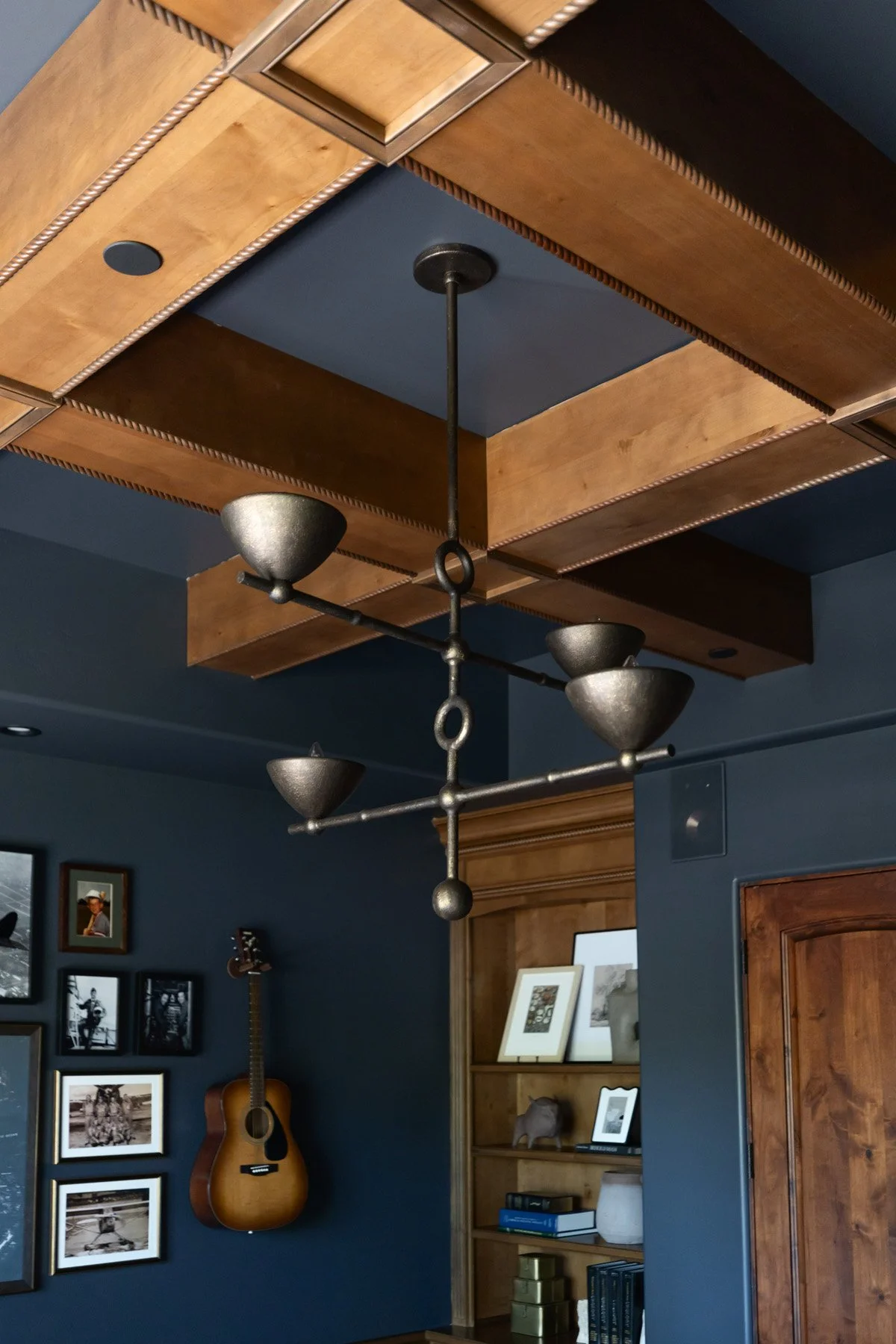 Overhead detail of dark coffered ceiling with exposed wood beams and sculptural pendant lights, with guitar on wall and framed artwork below