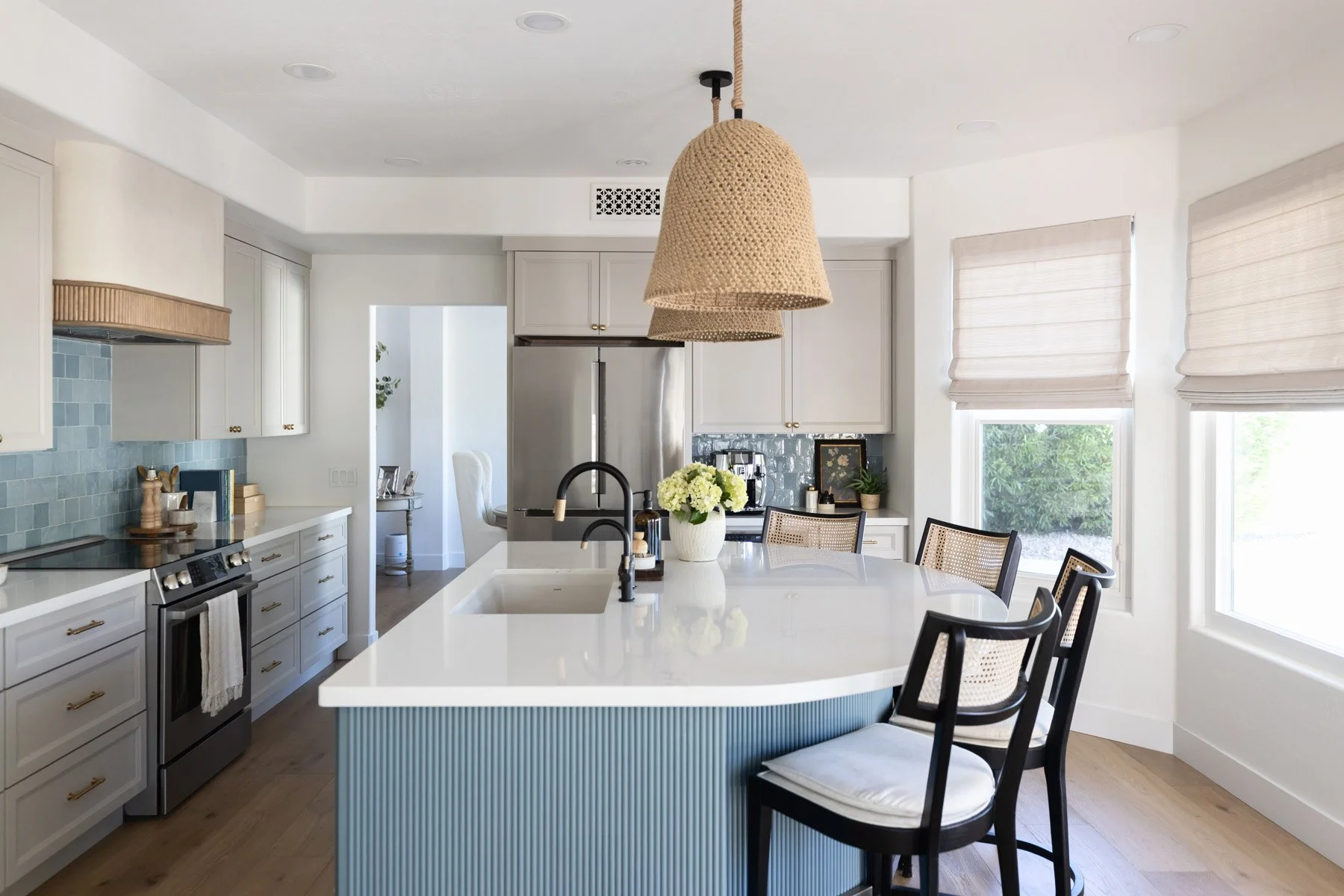  Kitchen with blue fluted island, white quartz countertop, black cane bar stools, woven pendant light, white shaker cabinetry, blue zellige tile backsplash, and linen roman blinds