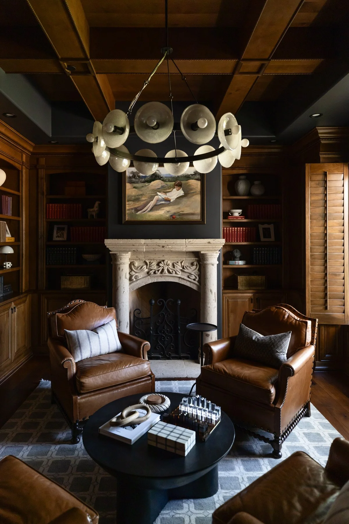 Dark study with leather armchairs, ornate stone fireplace, round black coffee table, sculptural pendant light, dark wood panelled ceiling, and built-in bookshelves