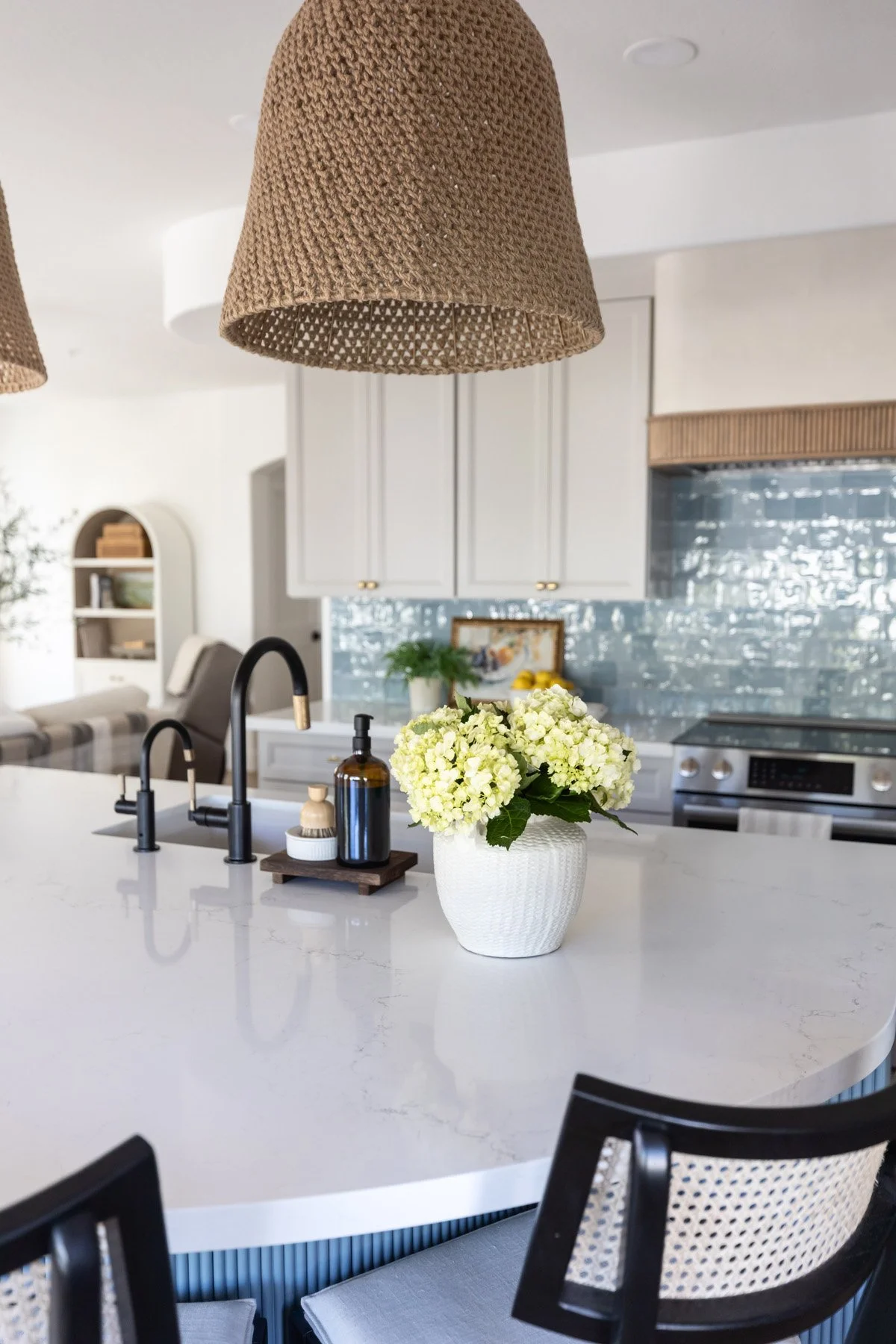Kitchen island detail with woven pendant light, white quartz countertop, black faucet, yellow floral arrangement, blue fluted island base, and cane bar stool