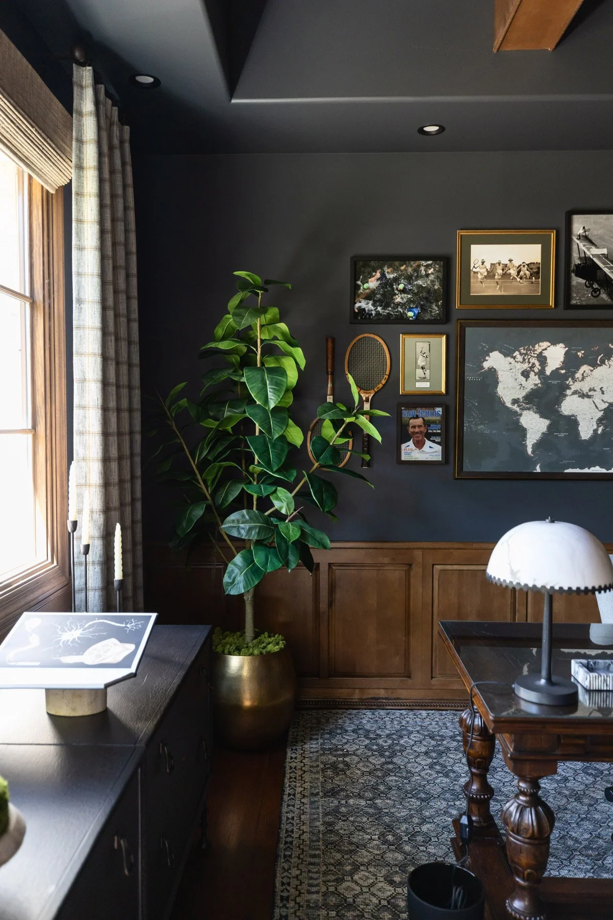Dark home office with navy walls, dark wood wainscoting, writing desk, white table lamp, world map artwork, framed photo gallery, potted fiddle leaf fig, and patterned rug