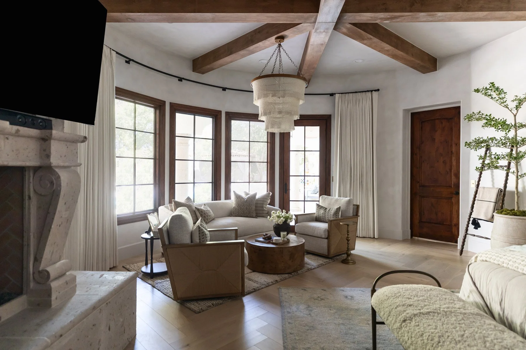 Primary suite with white sofas, stone fireplace, French windows with linen drapery, dark wood door, potted tree, and woven fringe pendant light