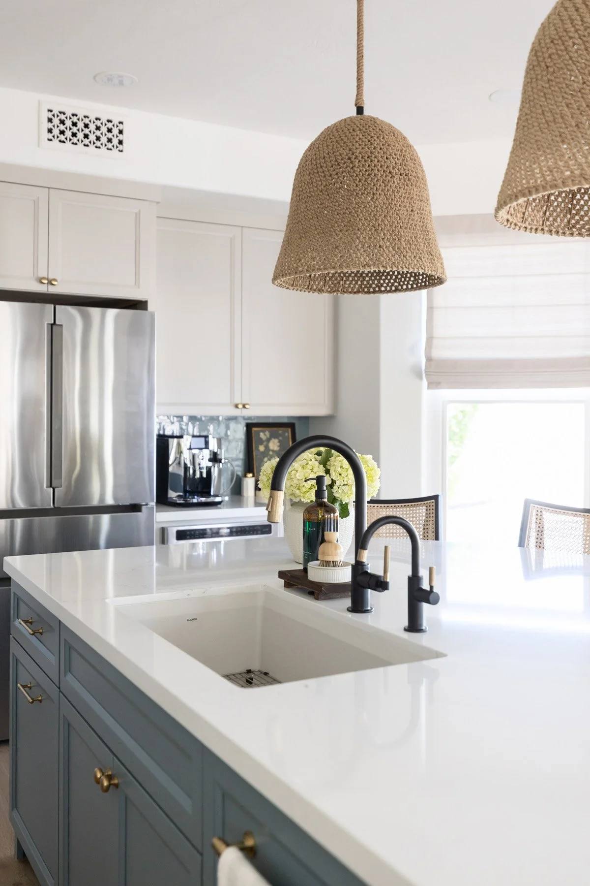Kitchen island sink detail with white quartz countertop, black faucet, blue fluted island base, woven pendant light, and stainless steel fridge