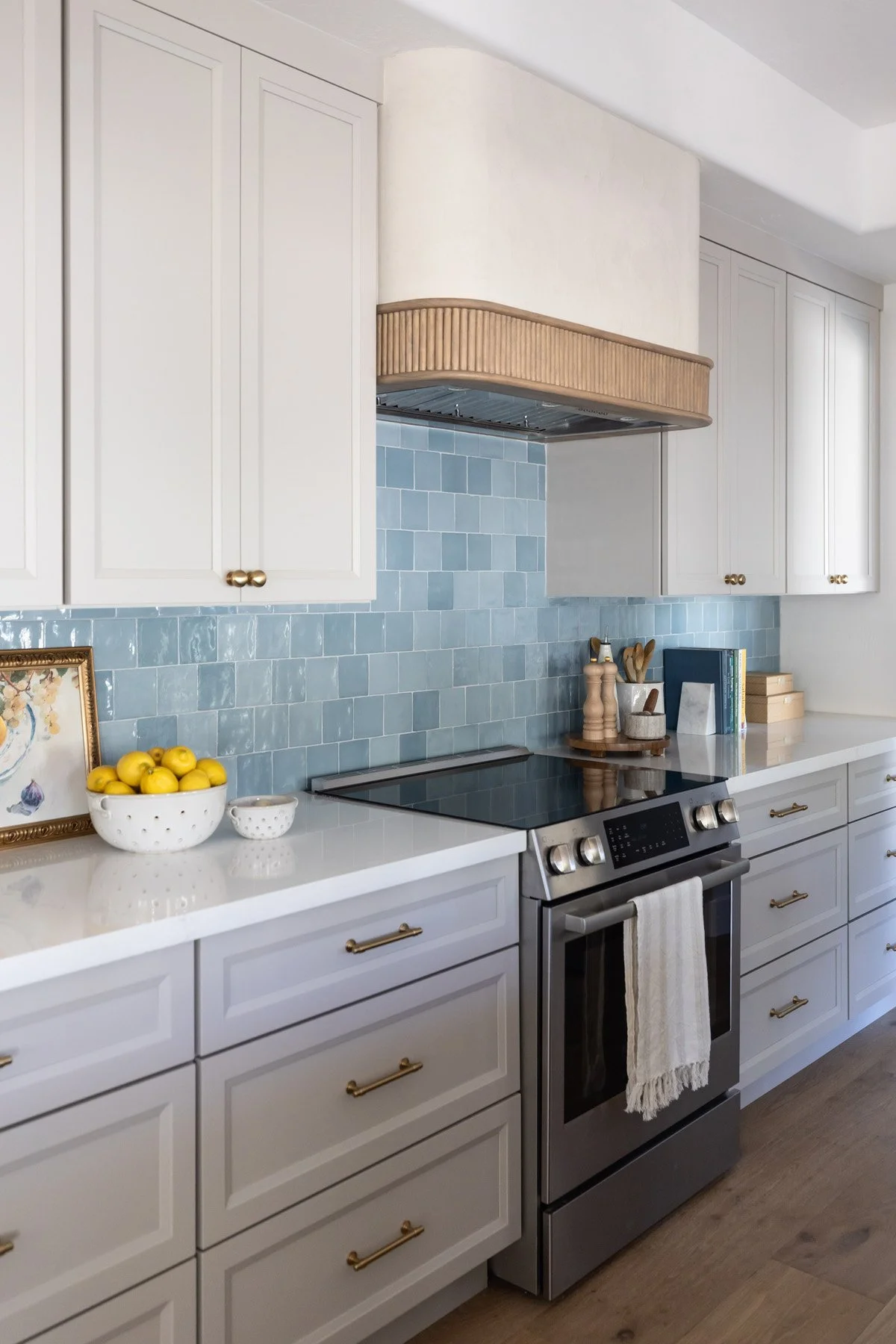 Kitchen range wall with white shaker cabinetry, brass hardware, woven range hood, blue zellige tile backsplash, stainless steel range, and bowl of lemons on counter