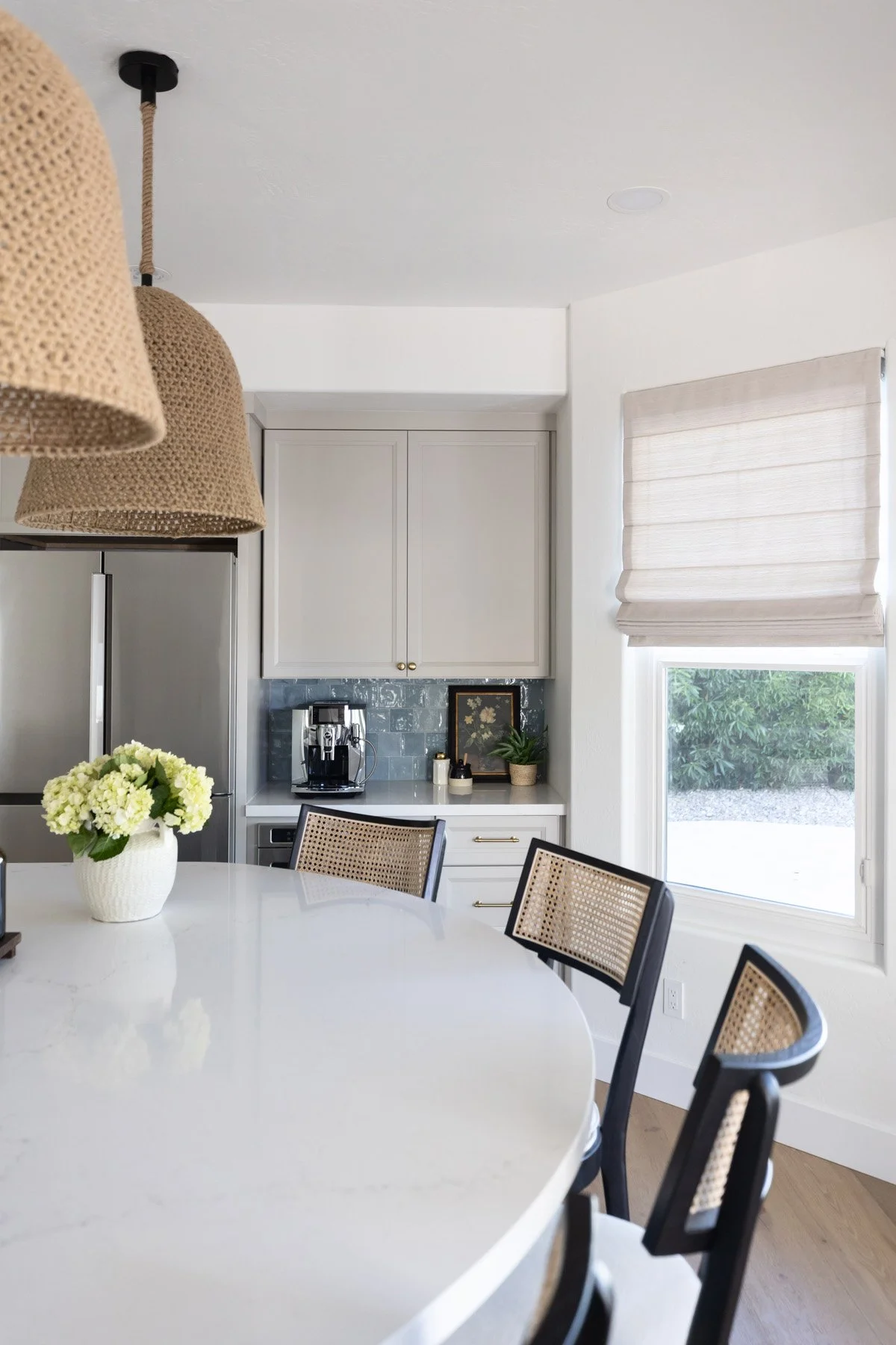 Bright coastal kitchen dining area with round white island, cane dining chairs, yellow floral arrangement, woven pendant light, linen roman blinds, and stainless steel fridge