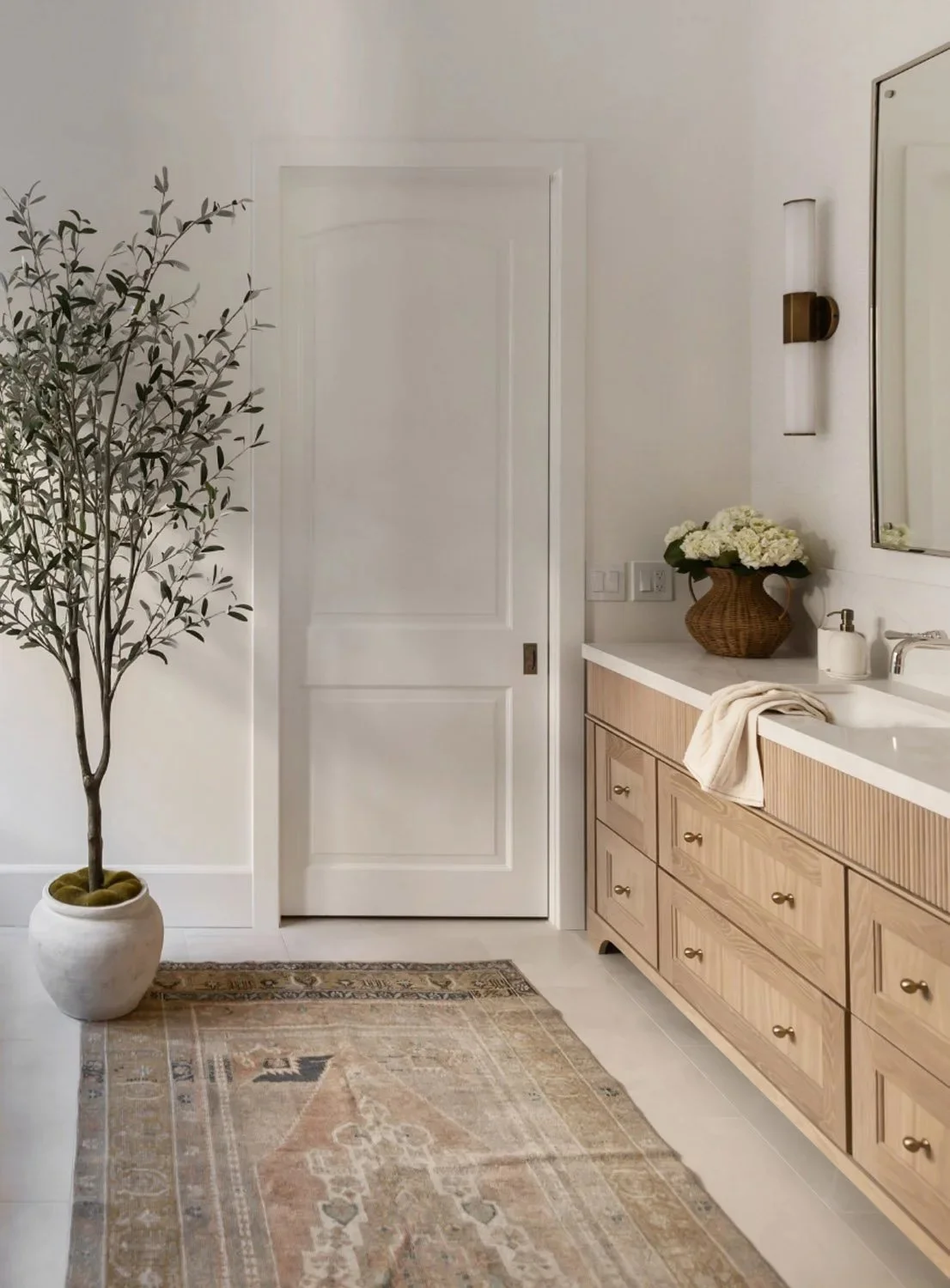 Light oak fluted vanity with potted olive tree and white panelled door, vintage-style rug, and brass wall sconce