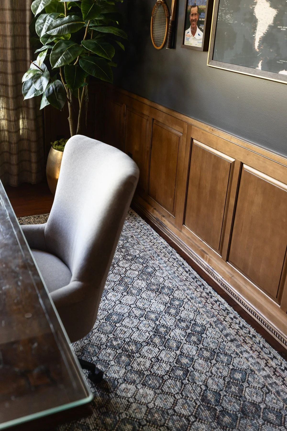 Grey upholstered chair on patterned blue rug beside dark wood wainscoting, with potted tree and framed portrait beyond