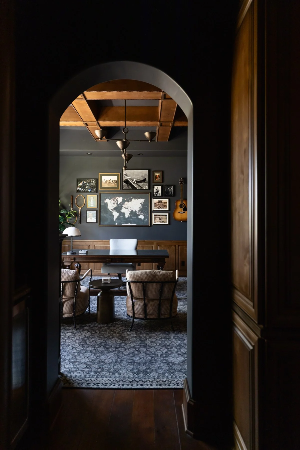 Dark arched doorway leading into a study with dark wood panelling, built-in bookshelves, world map artwork, and patterned rug beyond