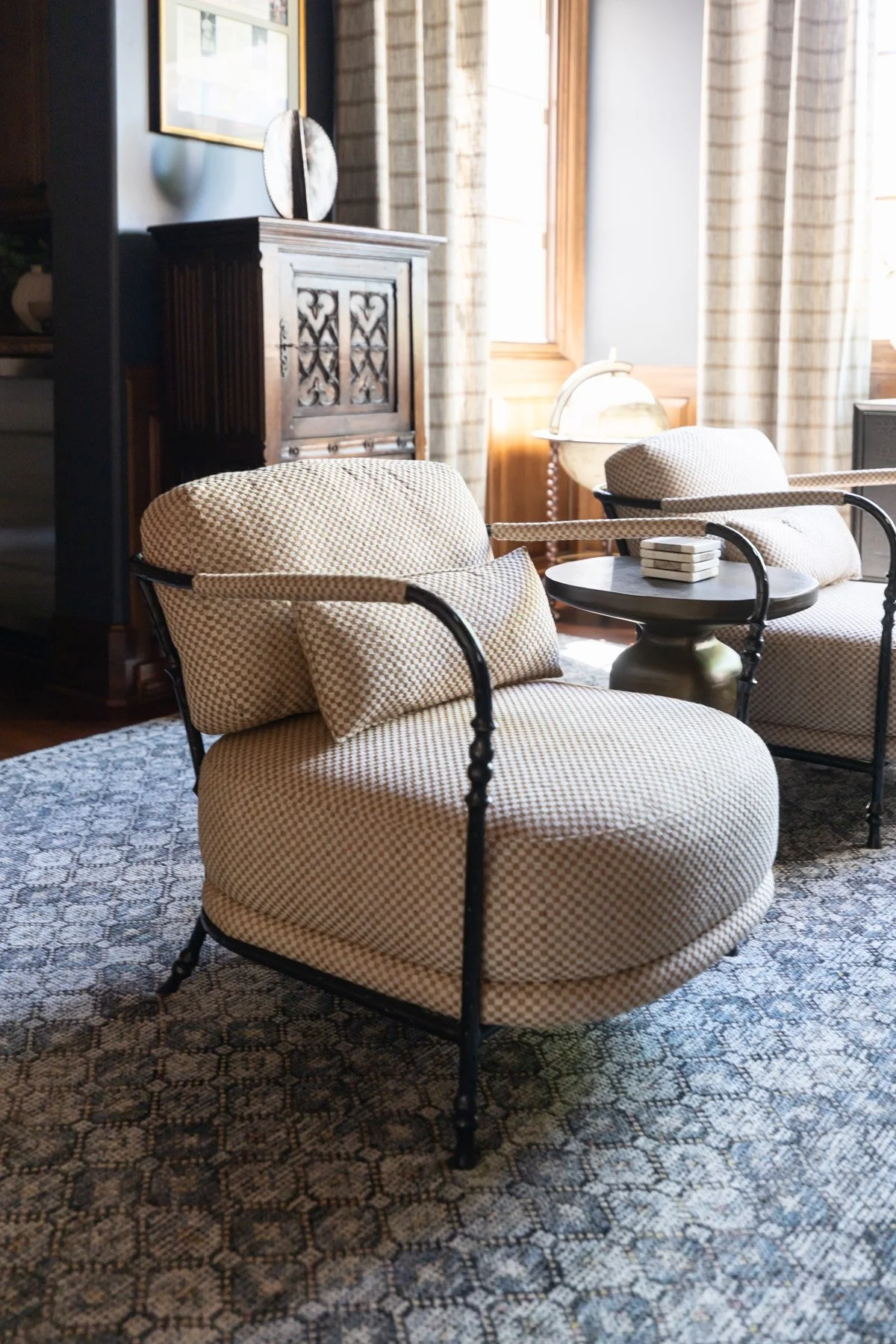Woven upholstered armchair with black iron frame on patterned blue rug with check drapery and dark wood cabinet beyond