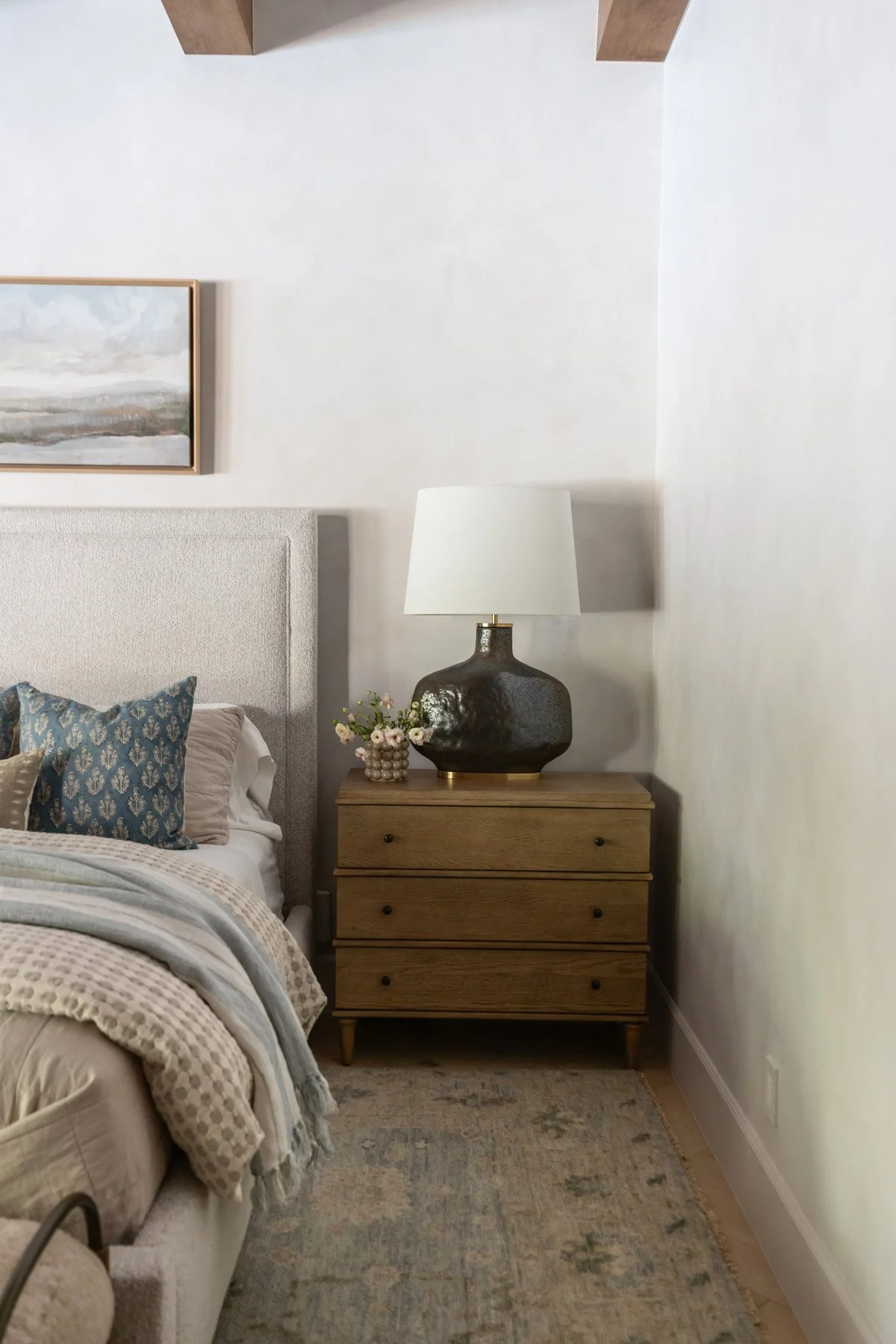 Primary bedroom corner with upholstered bedhead, blue accent pillows, oak bedside table, black ceramic lamp, and layered neutral bedding
