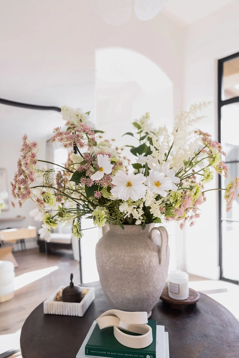 Large textured ceramic urn with handles filled with a lush arrangement of white cosmos, pink astrantia, and greenery on a dark round pedestal table, styled with a decorative tray and coffee table book