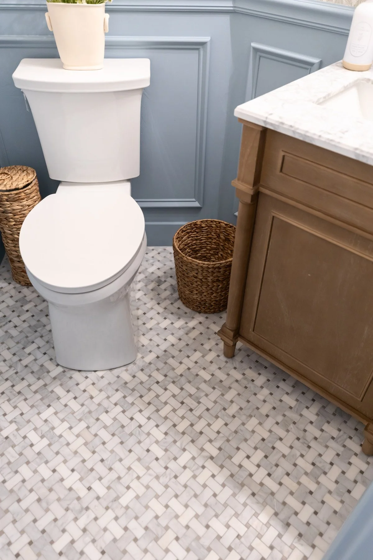 Powder room floor detail with patterned mosaic tile, blue painted skirting, dark wood vanity base, and woven basket