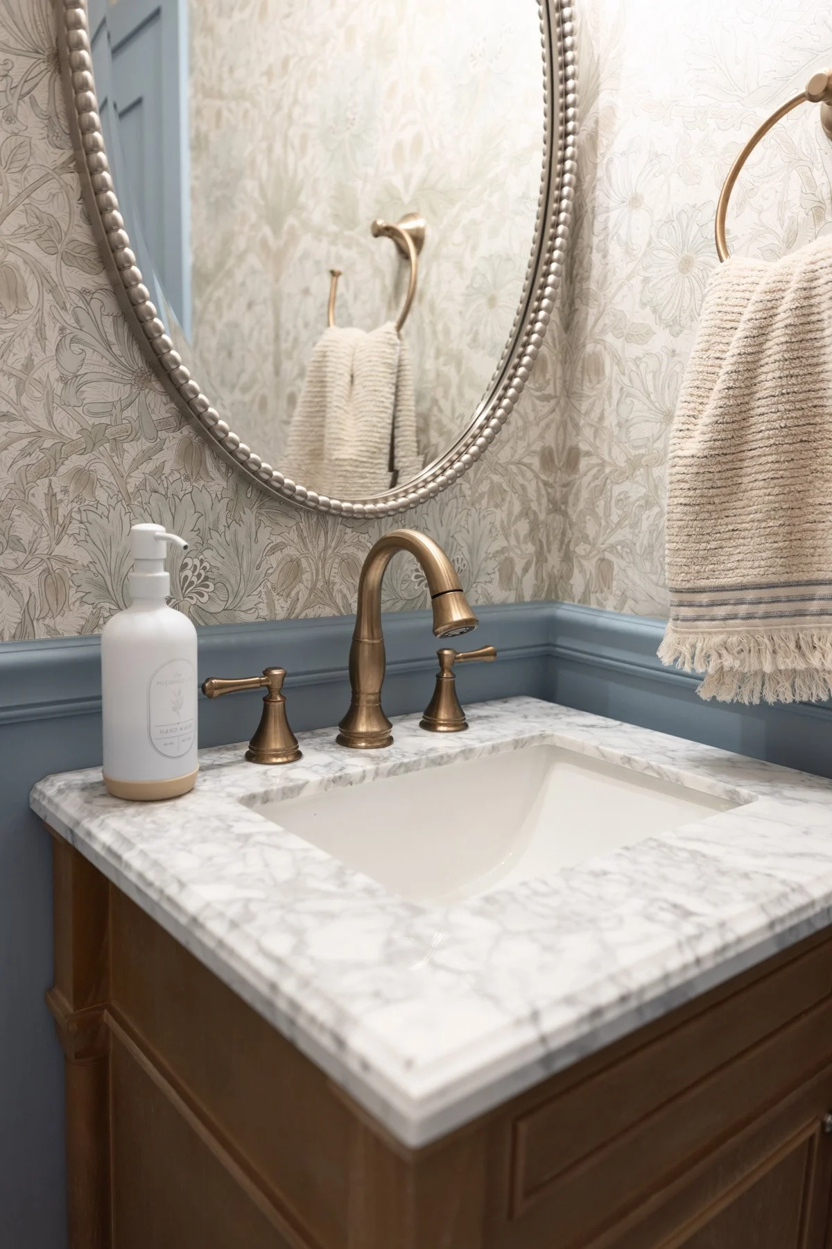 Powder room sink detail with marble countertop, brass widespread faucet, soap dispenser, oval mirror with beaded frame, and botanical wallpaper