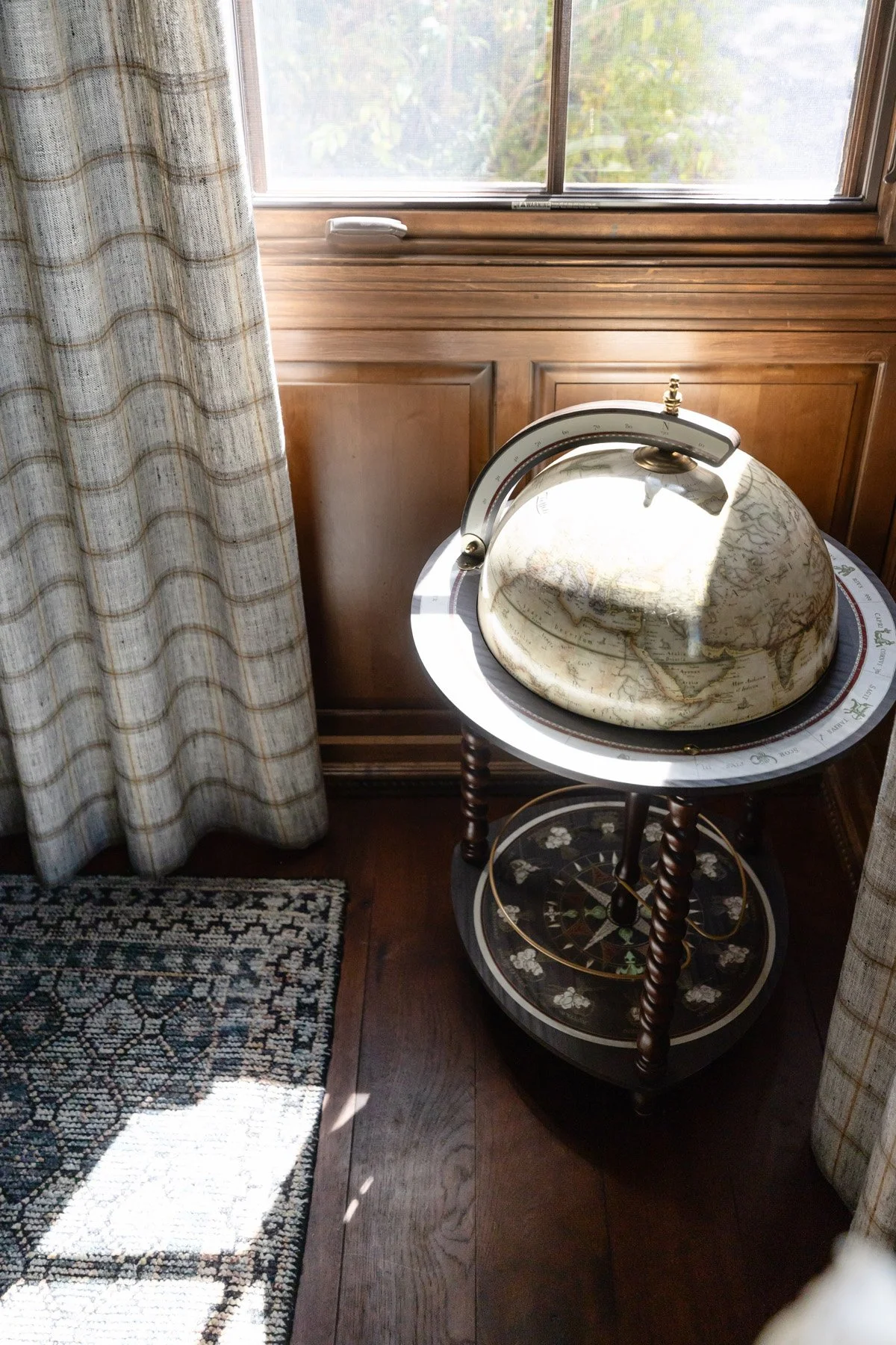 Vintage globe on dark wood stand beside window with check drapery and patterned rug below